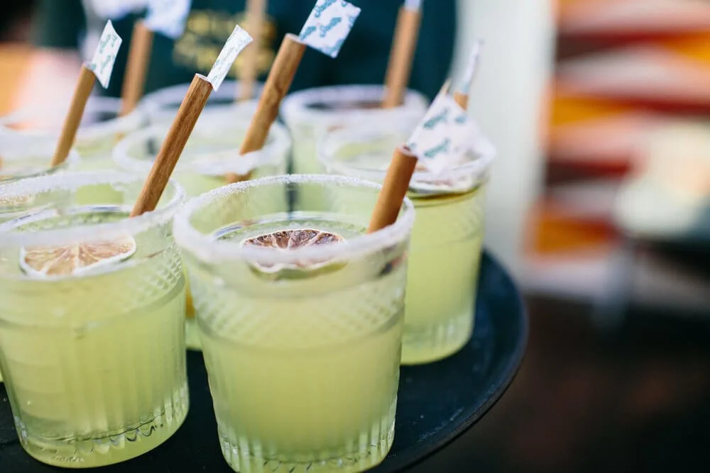 Several glasses of yellow lemonade with lemon slices, sugar rim, paper umbrella, and wooden stir stick, on a black tray.