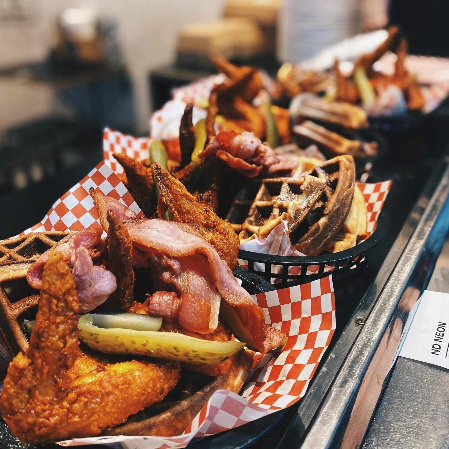 Basket of fried chicken, bacon, waffle, pickles, and waffle fries with checkered paper liner.