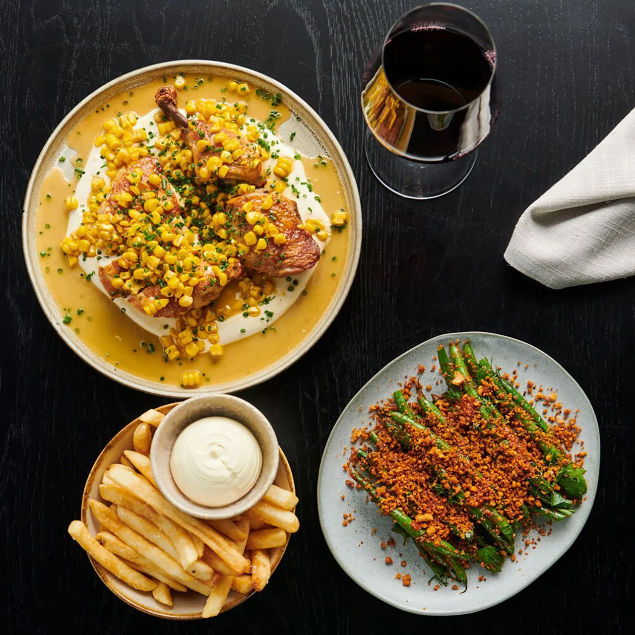 Plate with fried chicken topped with corn and chives, side of French fries with mayonnaise, green beans with breadcrumbs, and a glass of red wine on a dark table.