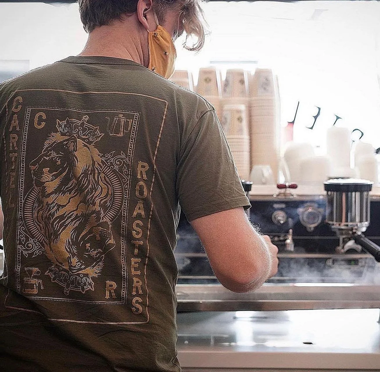 Back view of a person wearing a brown T-shirt with a lion and the words 'CARTIER ROASTERS' on the back, working at a coffee station with white cups and coffee brewing equipment.