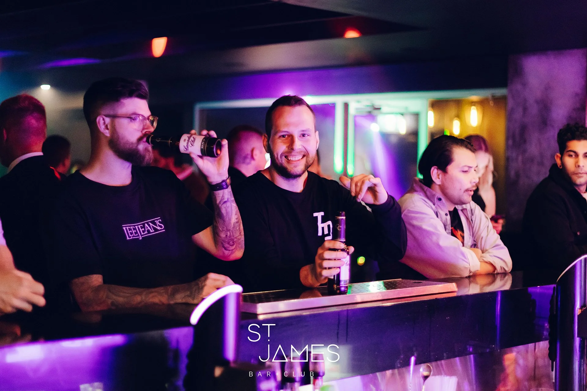 A group of men at a bar in a nightclub, with colorful neon lights illuminating the scene. Some are holding drinks, and they are standing at the bar counter, enjoying the nightlife.