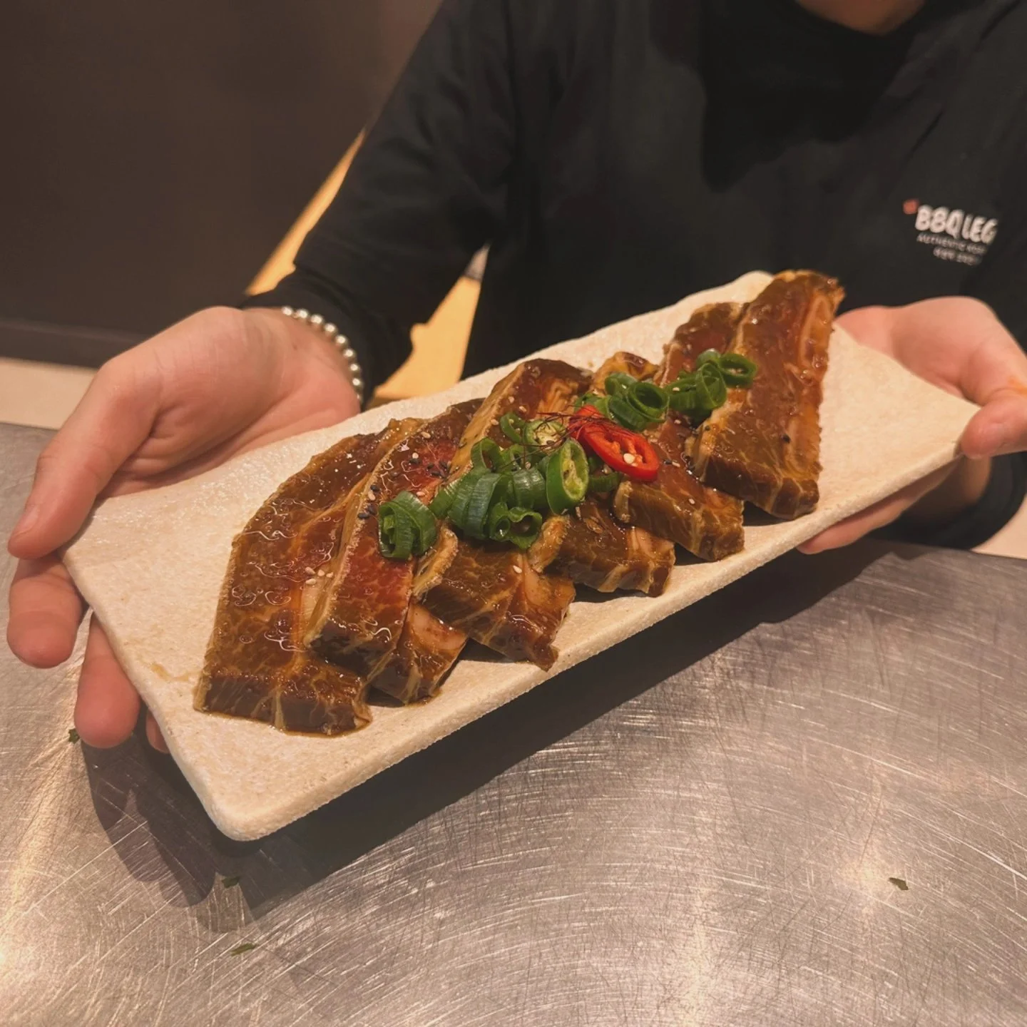 Person holding a rectangular ceramic plate with several pieces of cooked beef short ribs garnished with chopped green onions and sliced red chili peppers.