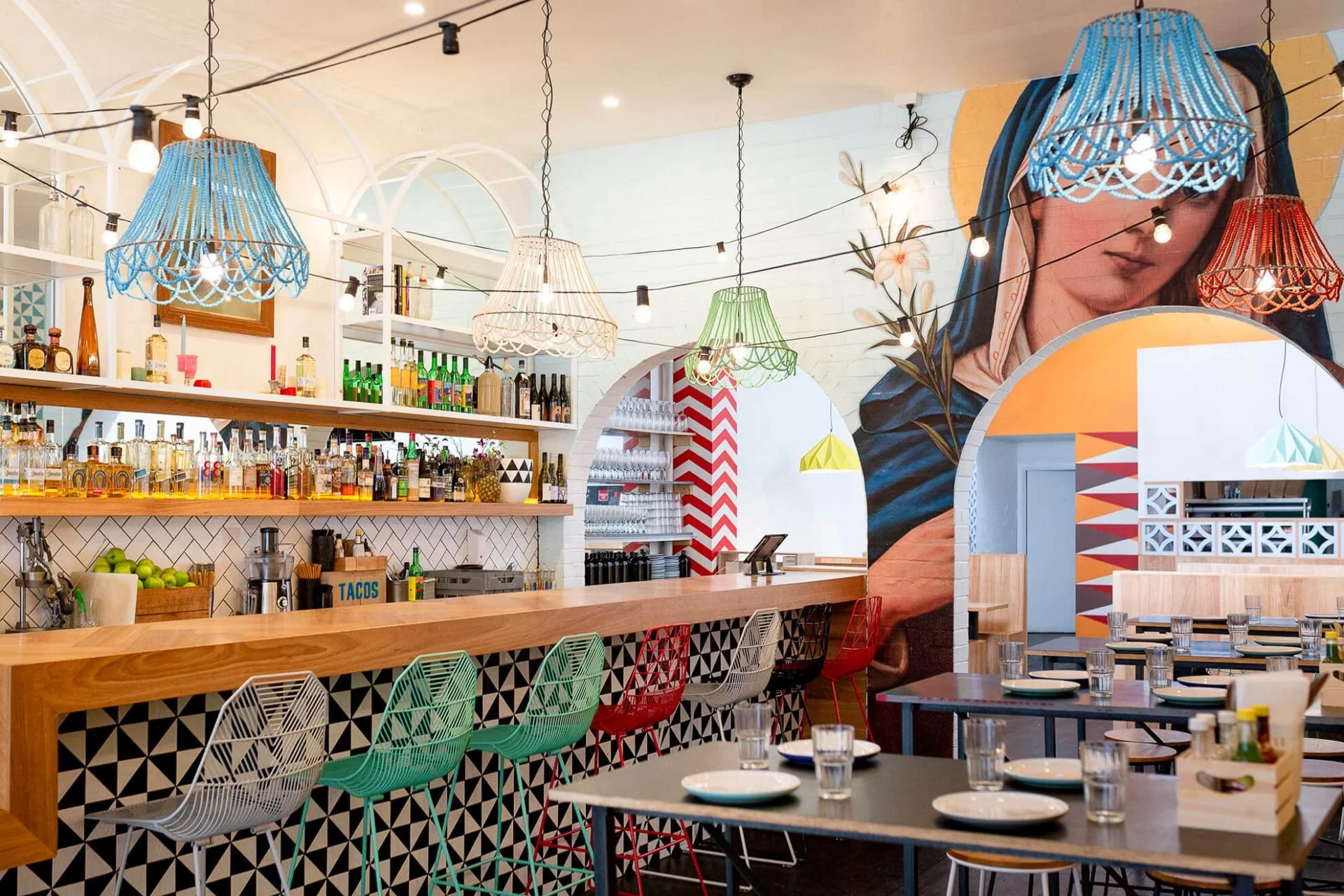 Interior of a colorful, modern restaurant with a bar, hanging pendant lights in various colors, and tables set with plates and glasses. The walls feature vibrant murals and decorative patterns.