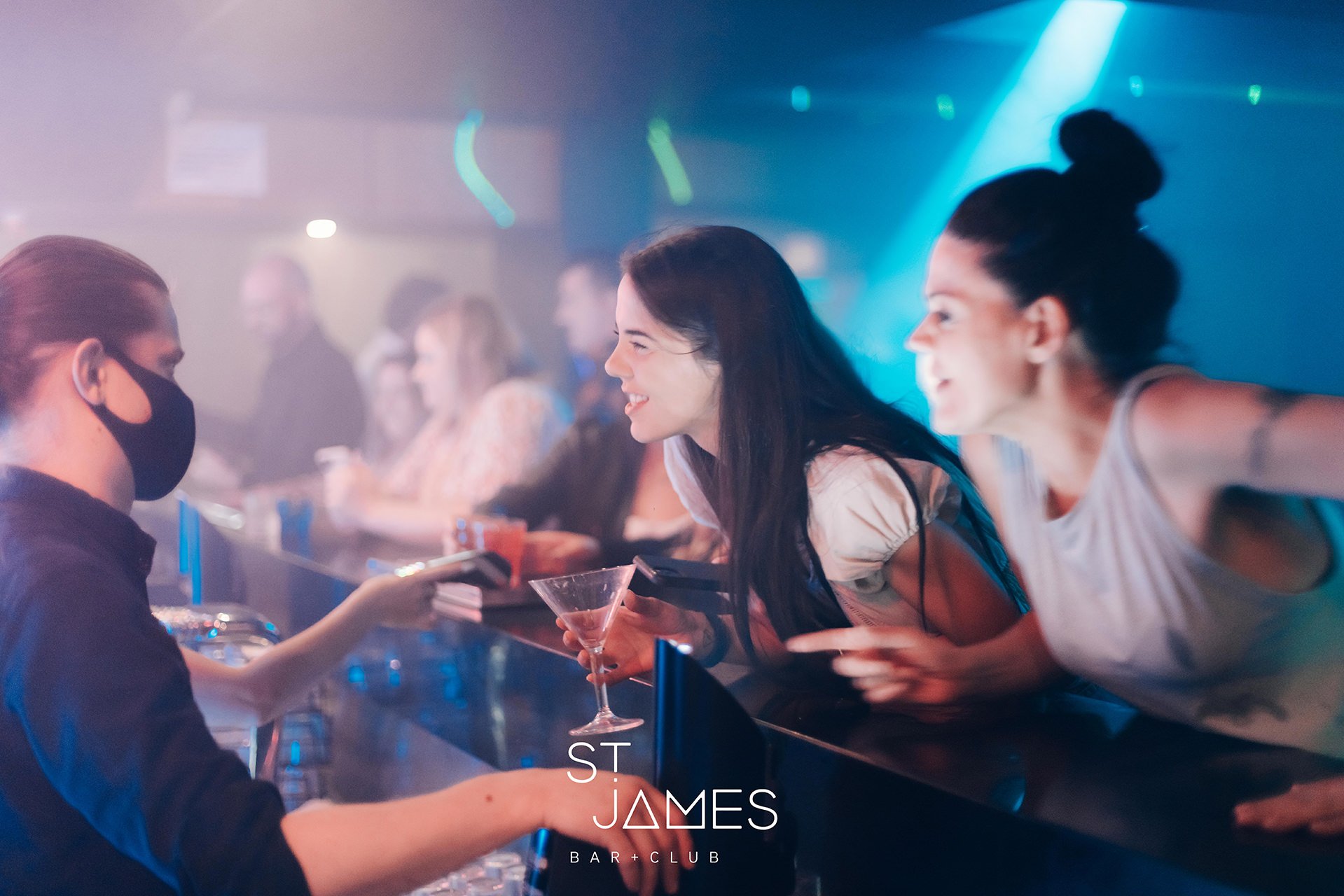 People at a bar lounge engaging with a bartender, with a woman smiling and holding a drink, illuminated by colorful nightclub lighting.