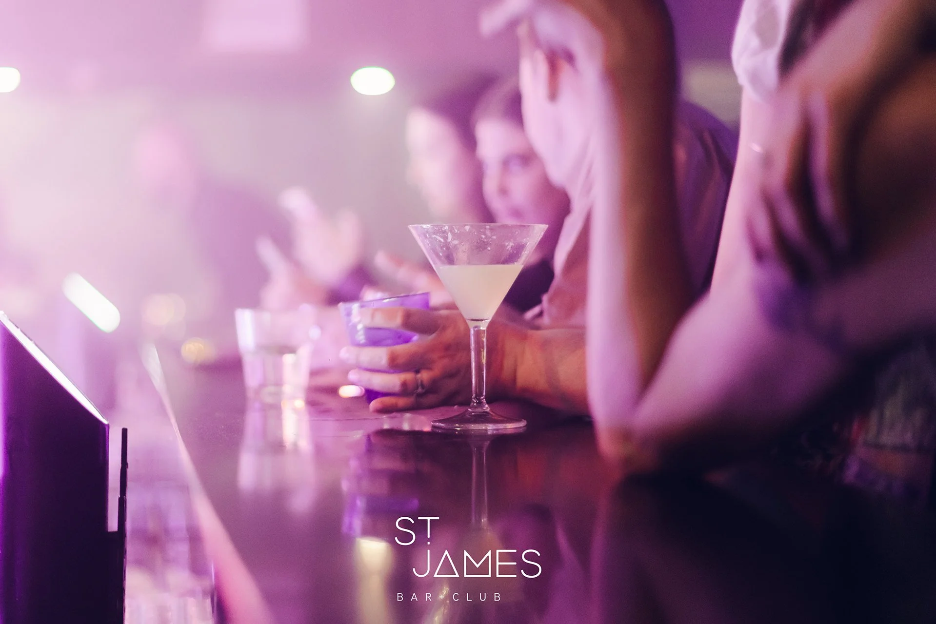 People sitting at a bar with drinks, illuminated by purple and pink lighting, at St James Bar & Club.