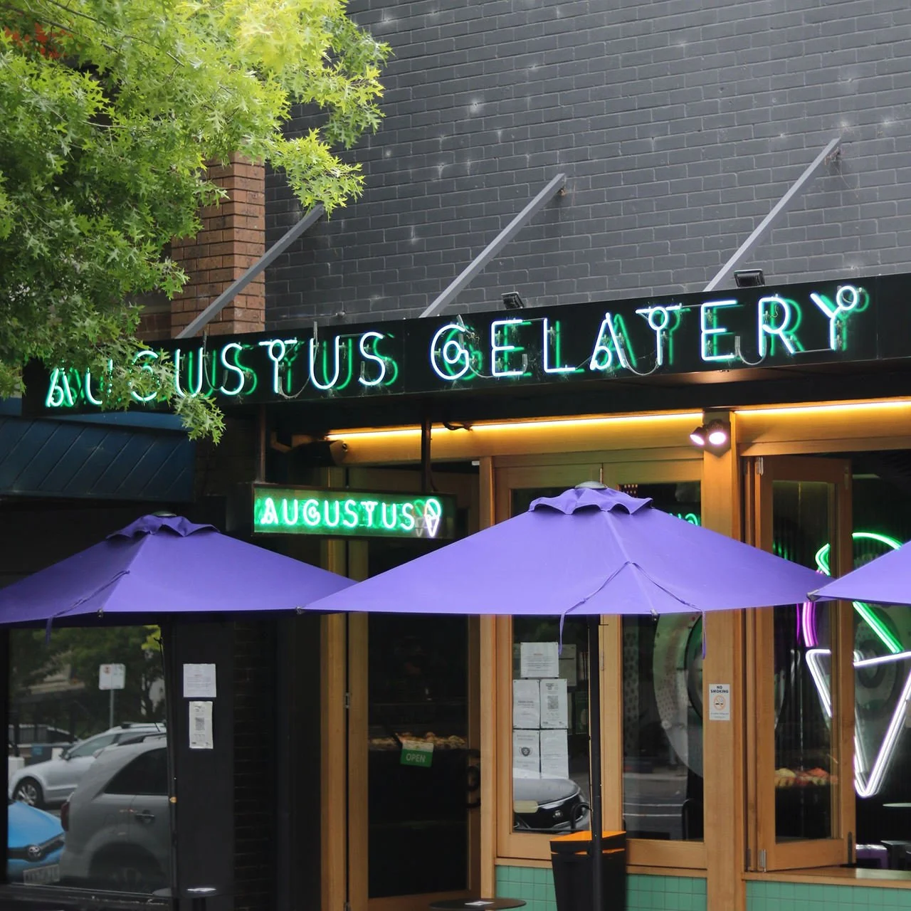 Neon sign reads 'AUGUSTUS GELATERY' outside a storefront with purple umbrellas and a tree beside the entrance.