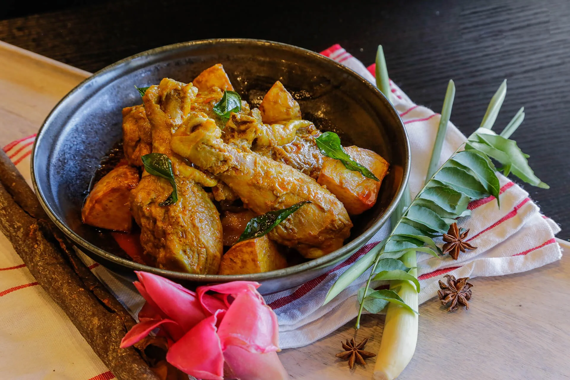 A bowl of curry with chicken and vegetables, garnished with green leaves, on a rustic wooden table with star anise, a pink flower, and leaves beside it.