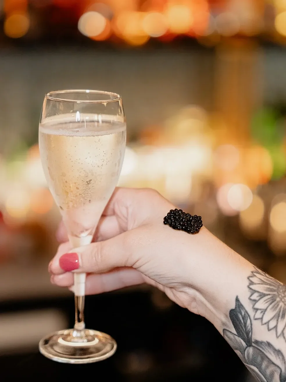 A hand with pink nail polish holding a glass of champagne with bubbles, and a black ring on the finger, against a blurred background of warm lights.
