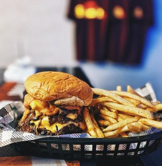 A cheeseburger with a beef patty, melted cheese, and a sandwich bun, served with French fries on a black plastic basket.