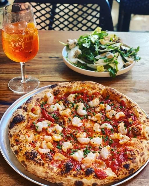 A shrimp pizza with cheese, tomato sauce, and herbs on a metallic plate, a leafy salad in a bowl, and a glass of iced tea on a wooden table.