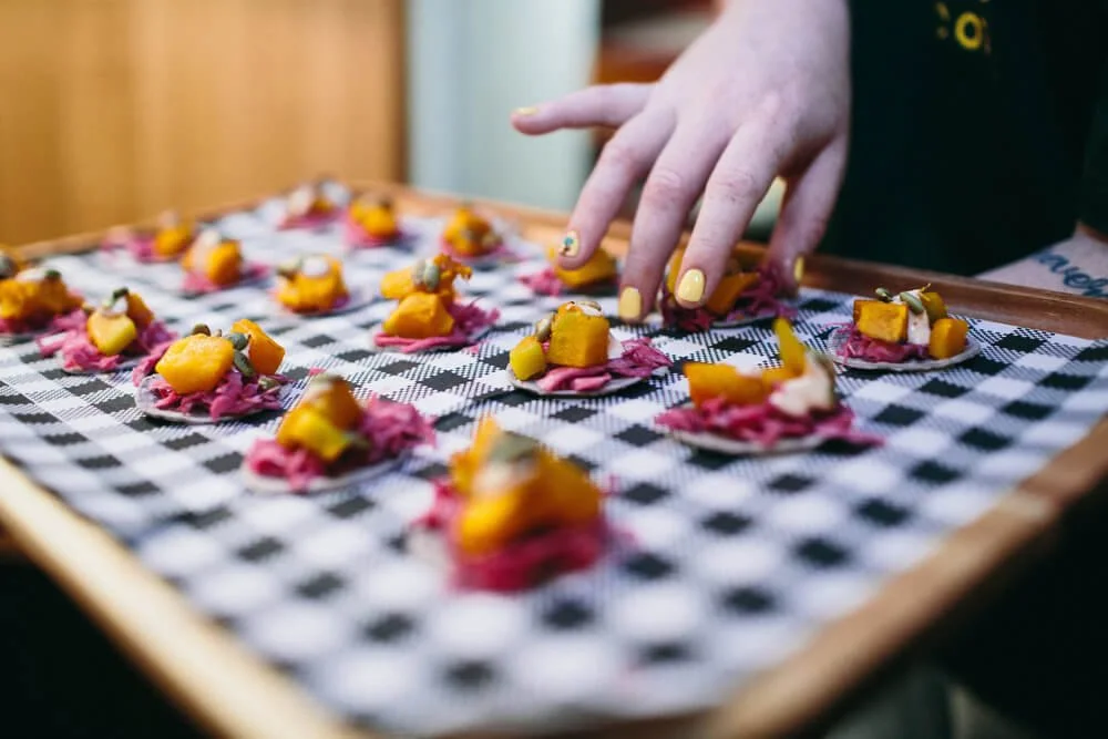 Person with yellow painted nails decorating mini appetizers with yellow cheese and pink shredded topping on a checkered tray