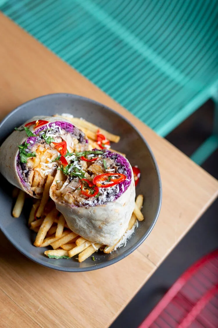 A bowl containing a wrap sushi roll with vegetables, red chili slices, and shredded cheese, placed on a bed of French fries on a wooden table.
