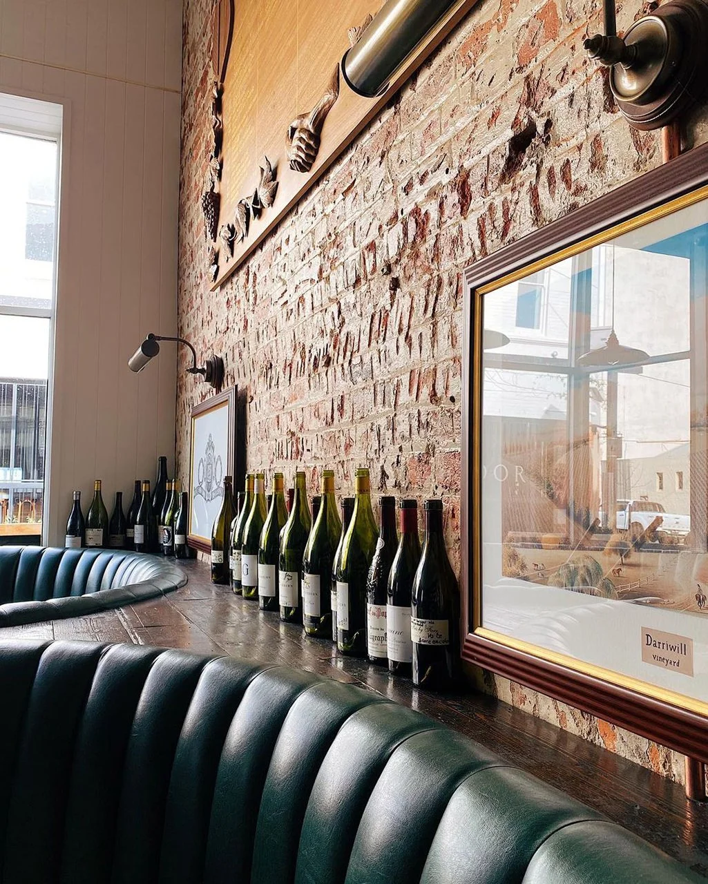 Interior of a restaurant or wine bar with a row of wine bottles on a wooden shelf against a brick wall, framed artwork, a large window, and green leather seating.