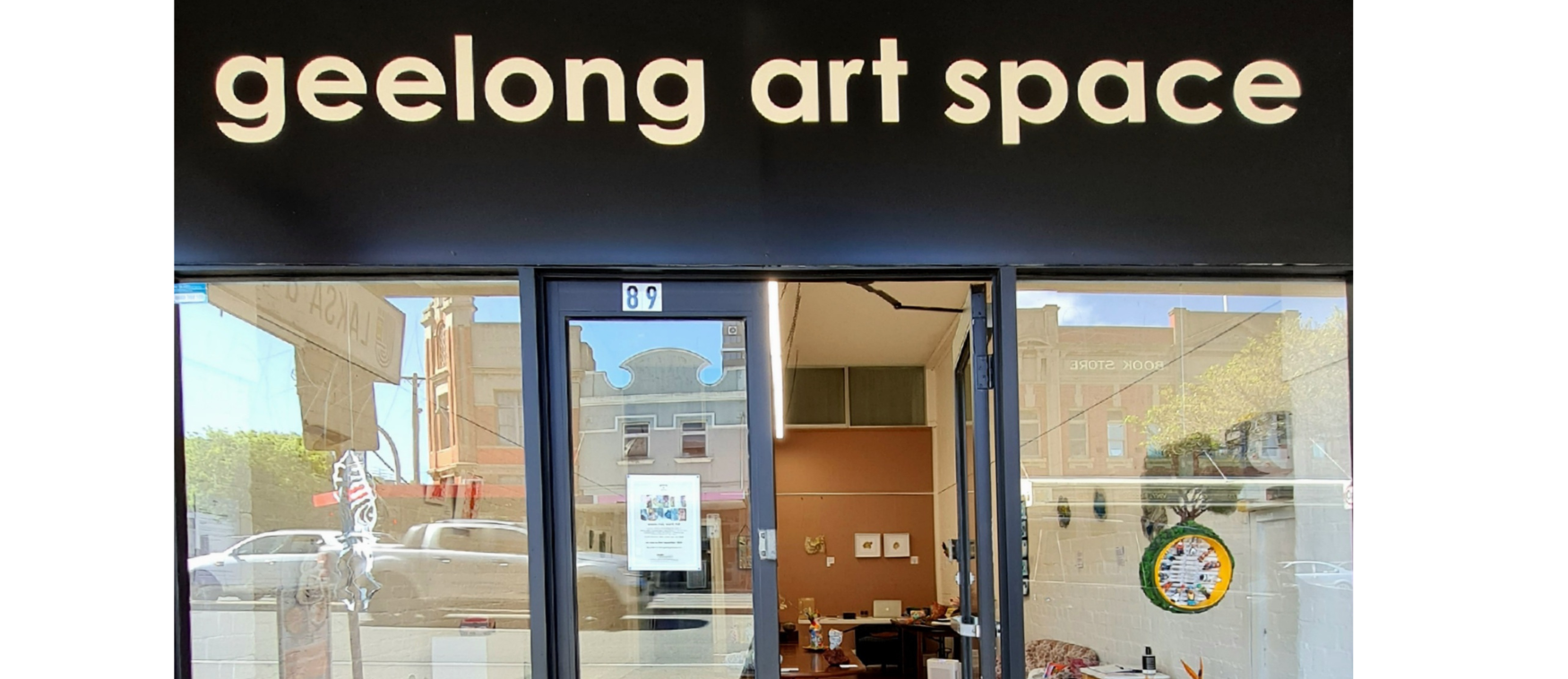 Exterior view of Geelong Art Space with glass door entrance, showing interior art gallery and reflection of buildings and street outside.