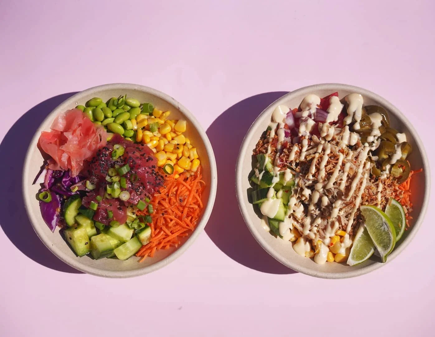 Two bowls of poke salad with vegetables and toppings, including jicama, edamame, corn, shredded carrots, cucumbers, purple cabbage, and pickled ginger, served on pink background.