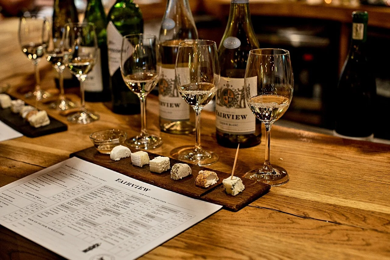Wine glasses filled with white wine, wine bottles, cheese samples on a wooden board, and a wine tasting menu on a wooden table.