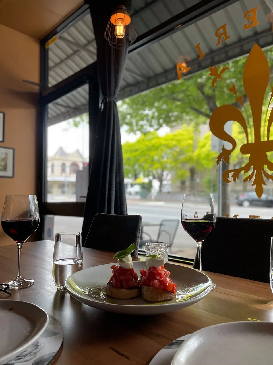 A plate of bruschetta topped with chopped tomatoes and garnished with basil leaves, with two glasses of red wine and a glass of water on a wooden table in a restaurant with large windows and outdoor views.