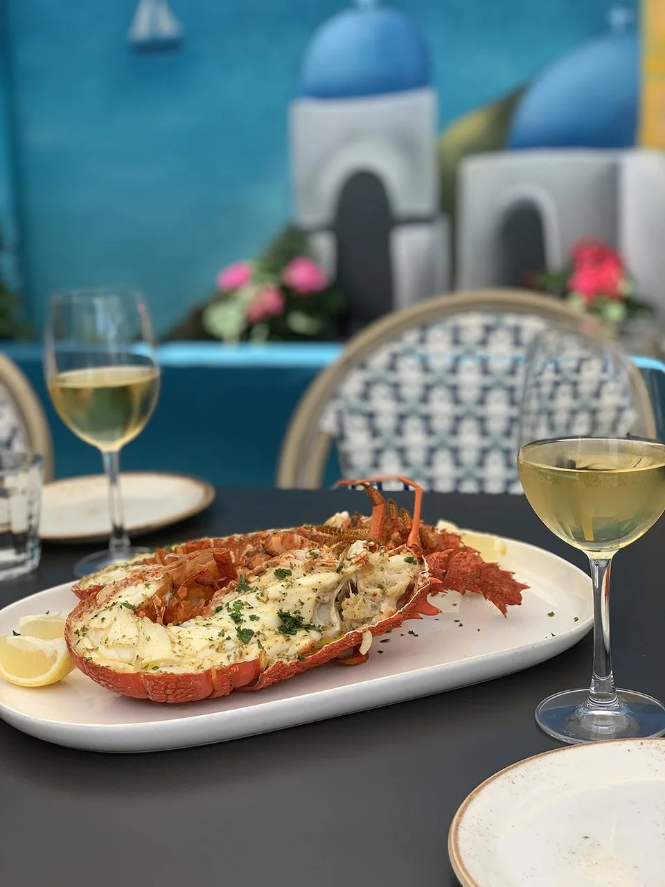 Lobster tail served on a white platter with lemon wedges, accompanied by two glasses of white wine, set on a dark table with a blurred colorful background.