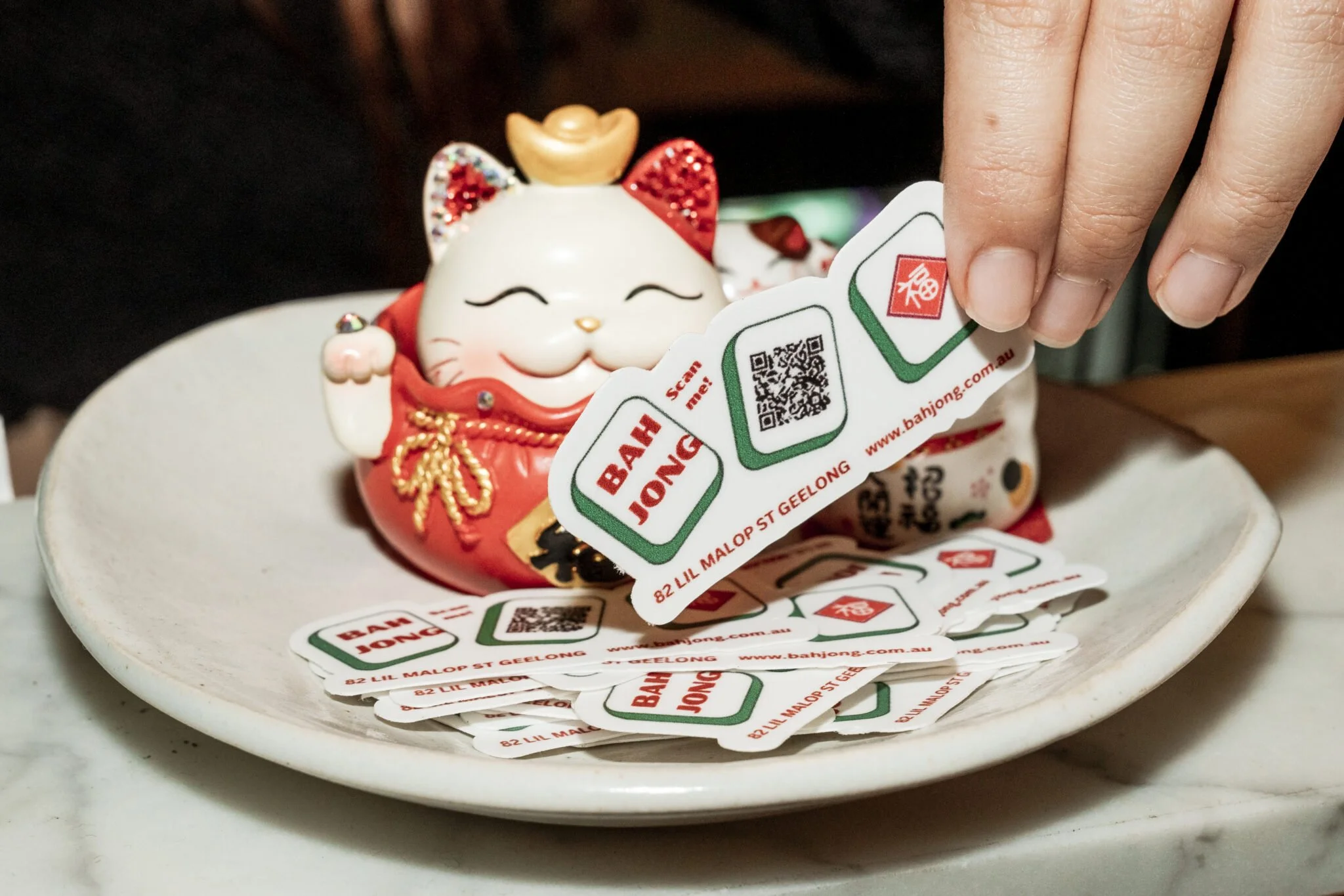 A ceramic Maneki-neko or lucky cat figurine in white with red accents, gold embellishments, and a smiling face, sitting on a white ceramic dish. A person's hand is holding a strip of Bah JONG coupons with QR codes and website information.