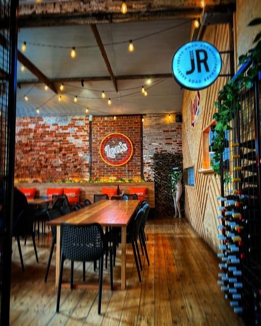 Interior of a restaurant or cafe with wooden tables and black chairs, brick and wood wall decor, neon signs for Louis's and JR, string lights hanging from the ceiling, and a wine rack on the right side.