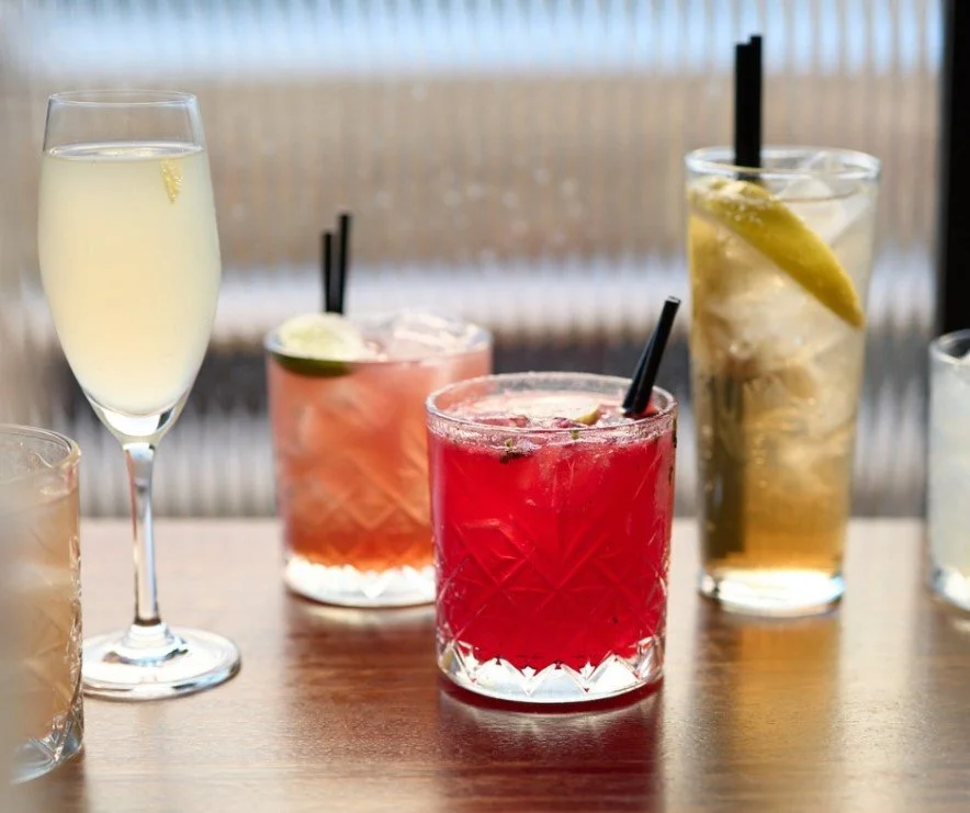Five different cocktails with garnishes on a wooden table, including champagne, fruity mixed drinks, and lemonade.