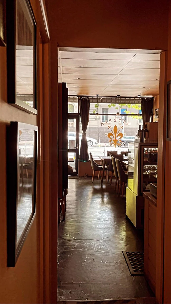 View of the interior of a cafe or restaurant with tables and chairs near a large front window with dark curtains, and a street scene outside.