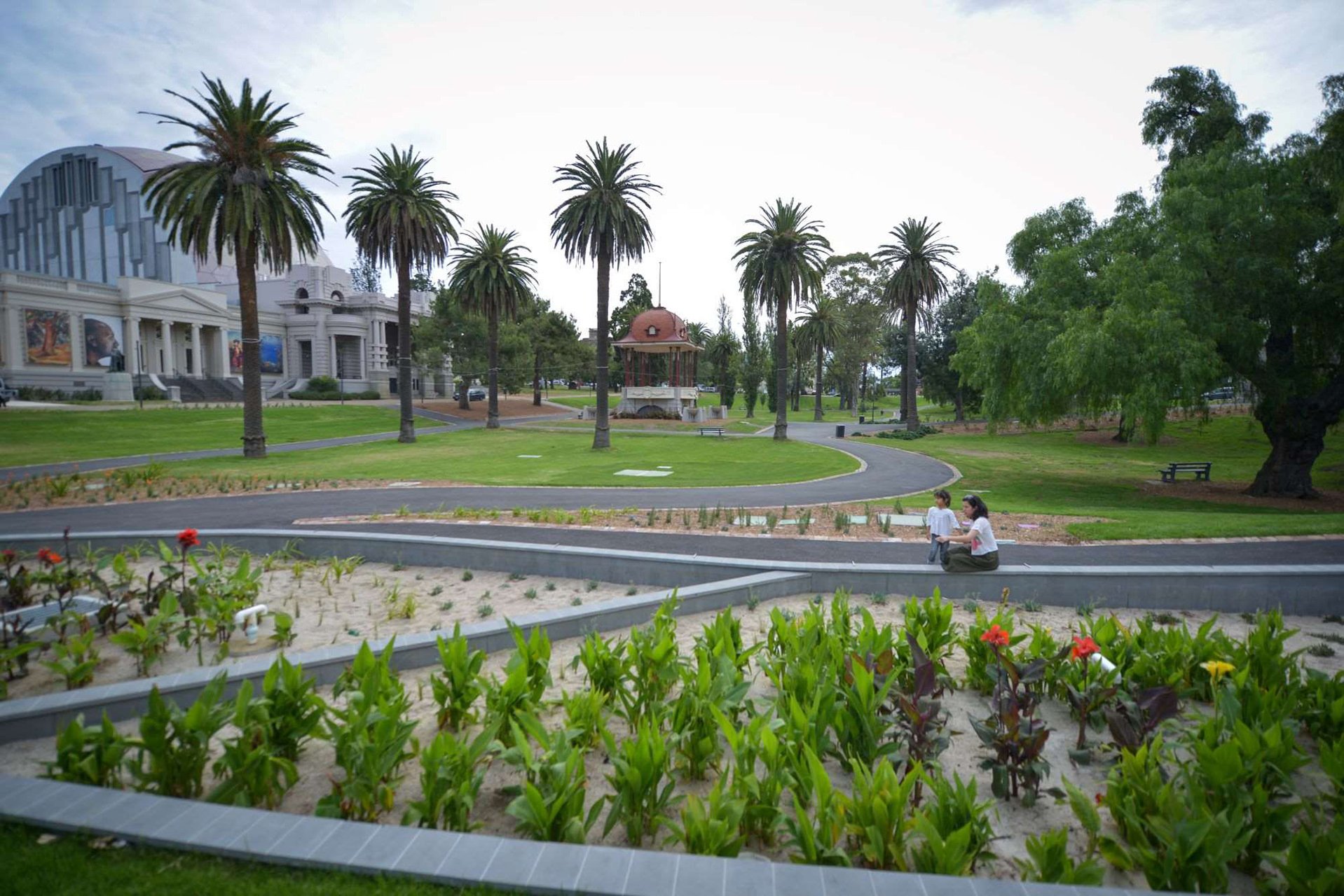 A park with palm trees, a small gazebo, and a modern building in the background. Two women sit on a low wall with a child nearby, and there are garden beds with plants and flowers in the foreground.