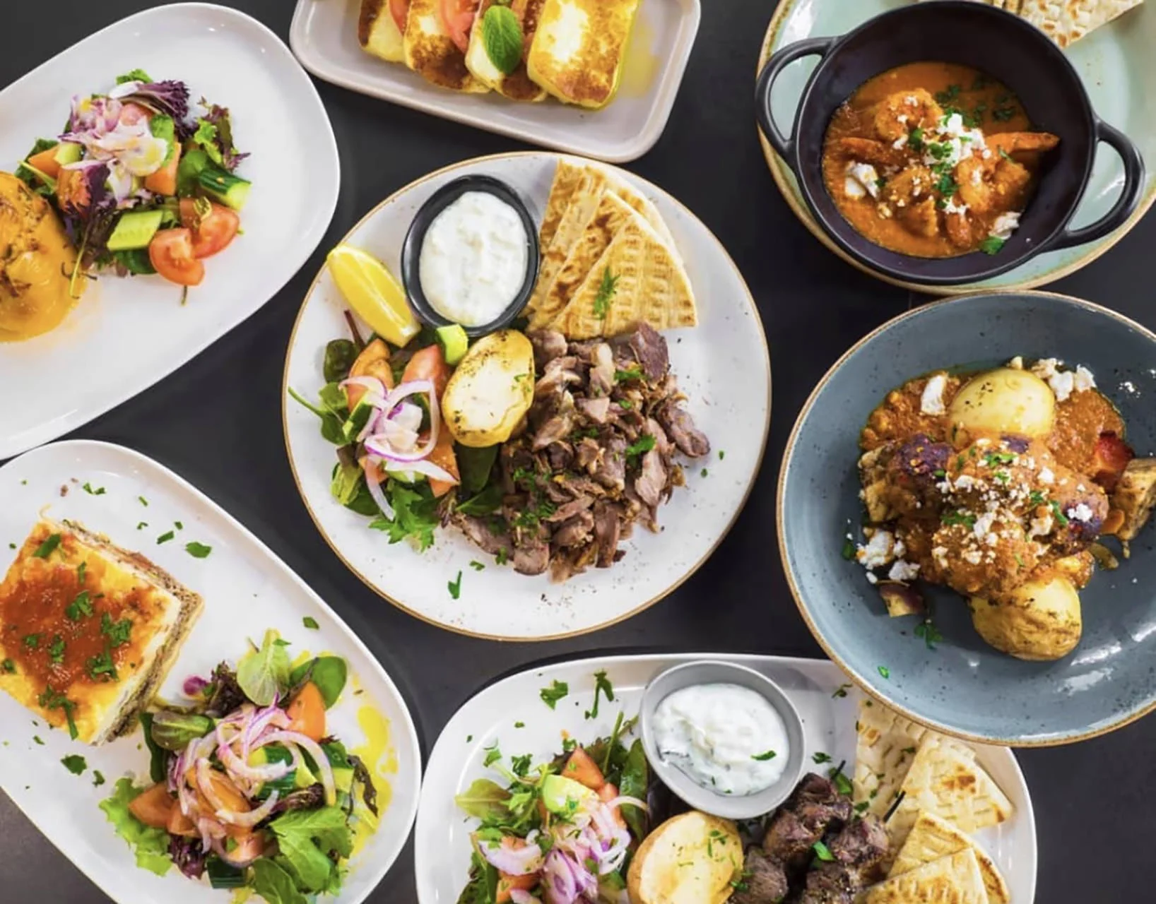 Assorted plates of Middle Eastern cuisine including salads, grilled meats, a bowl of stew, baked casserole, and flatbread, garnished with herbs.