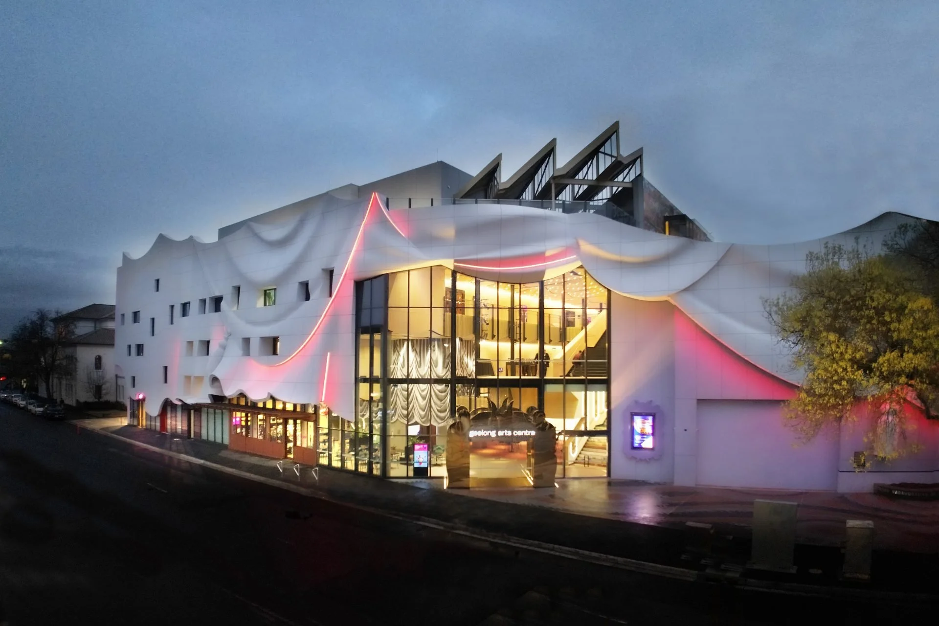 Nighttime view of the Geelong Arts Centre building with a distinctive white, wavy exterior and large glass facade, illuminated with pink and yellow lighting.