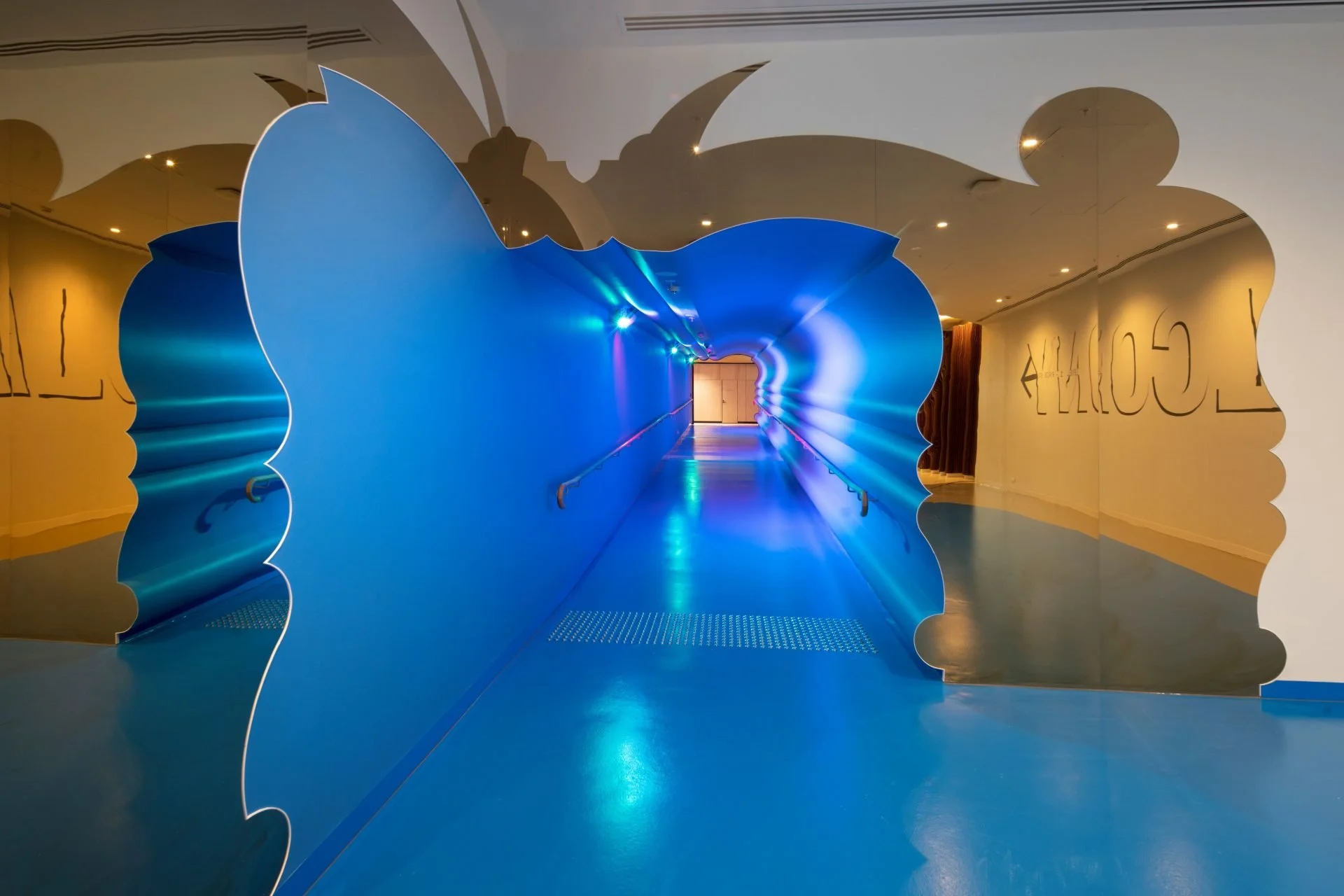 Art installation resembling a blue flower with a pathway through the center, mirror on the right reflecting the scene, and yellow walls with black writing in the background.