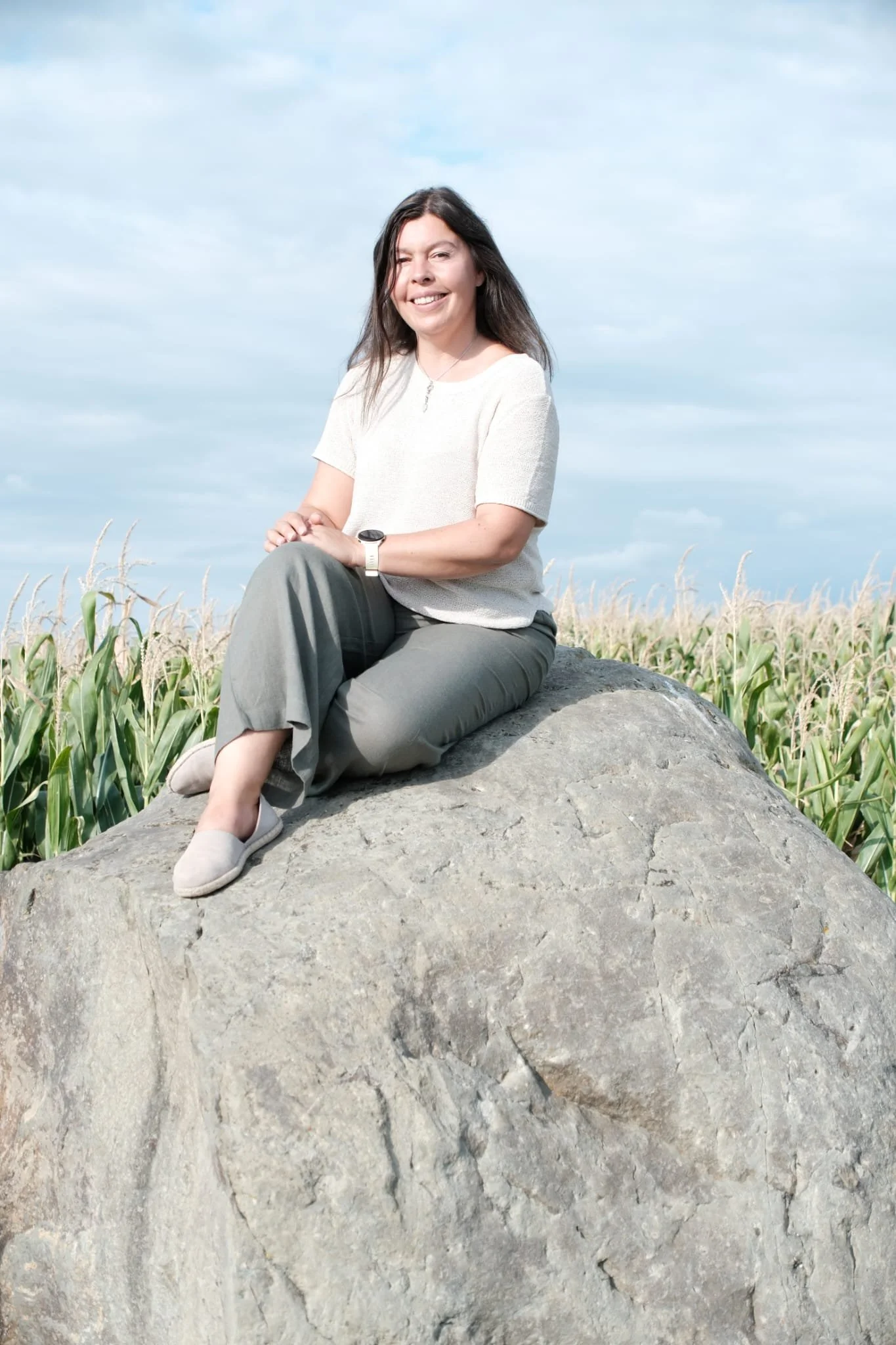 A woman with dark hair smiling, sitting on a large rock in a field of tall green plants under a partly cloudy sky.