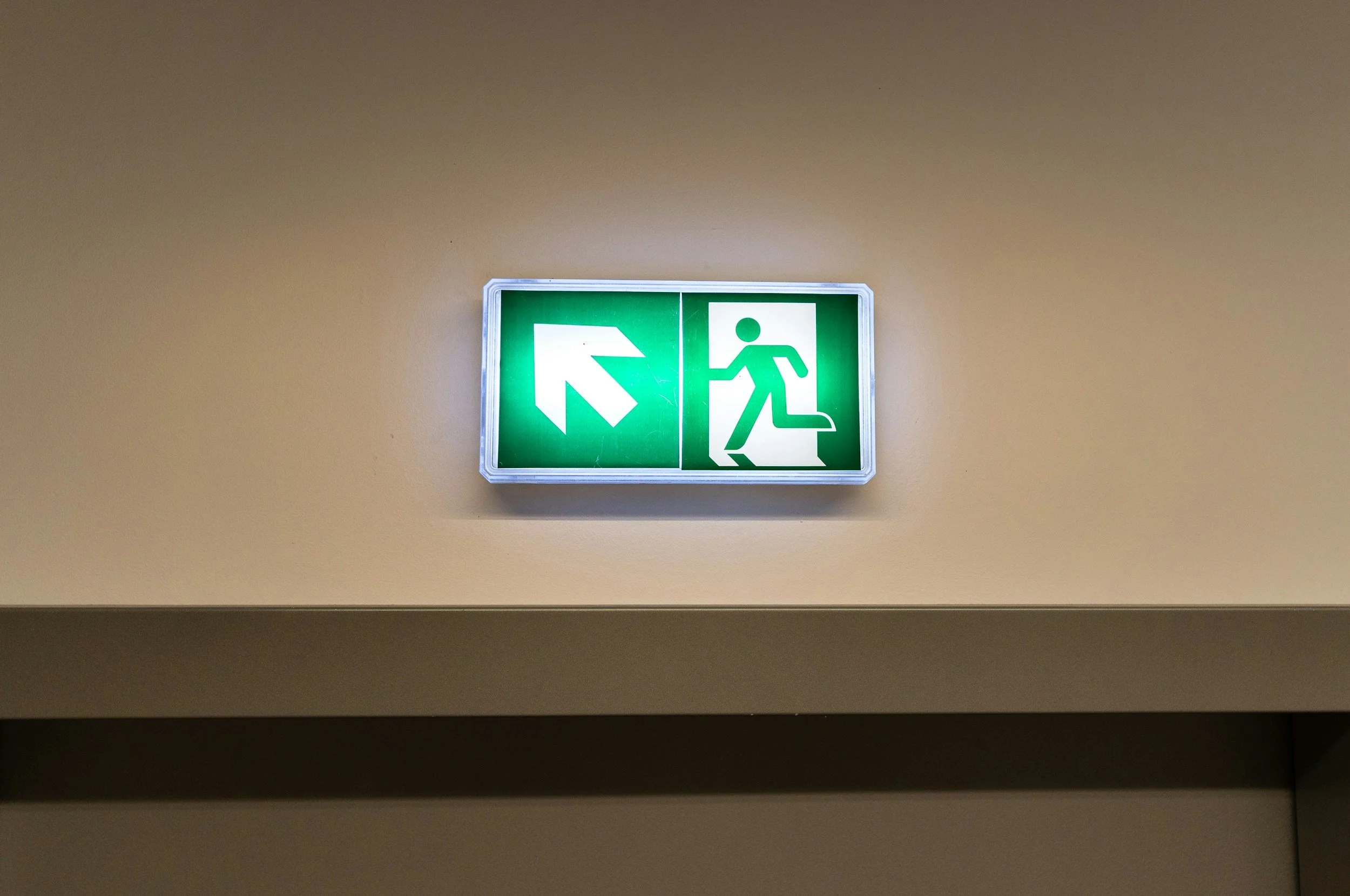 Green emergency exit sign with white arrow pointing left and a running figure climbing stairs on a beige wall.