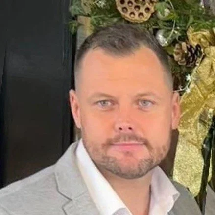 A man with short dark hair, light blue eyes, and a beard, wearing a light gray suit jacket and white shirt, standing indoors with a decorative floral arrangement in the background.