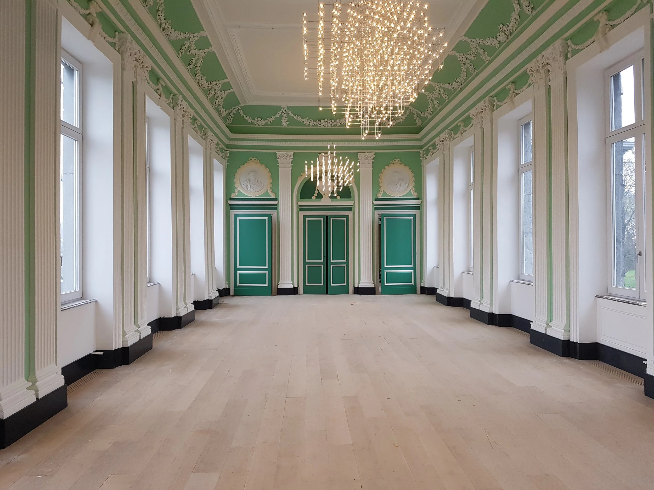 Salle ornée avec des murs de couleur vert et blanc, grands fenêtres, plafond décoratif avec des moulures, et un chandelier moderne suspendu au plafond.