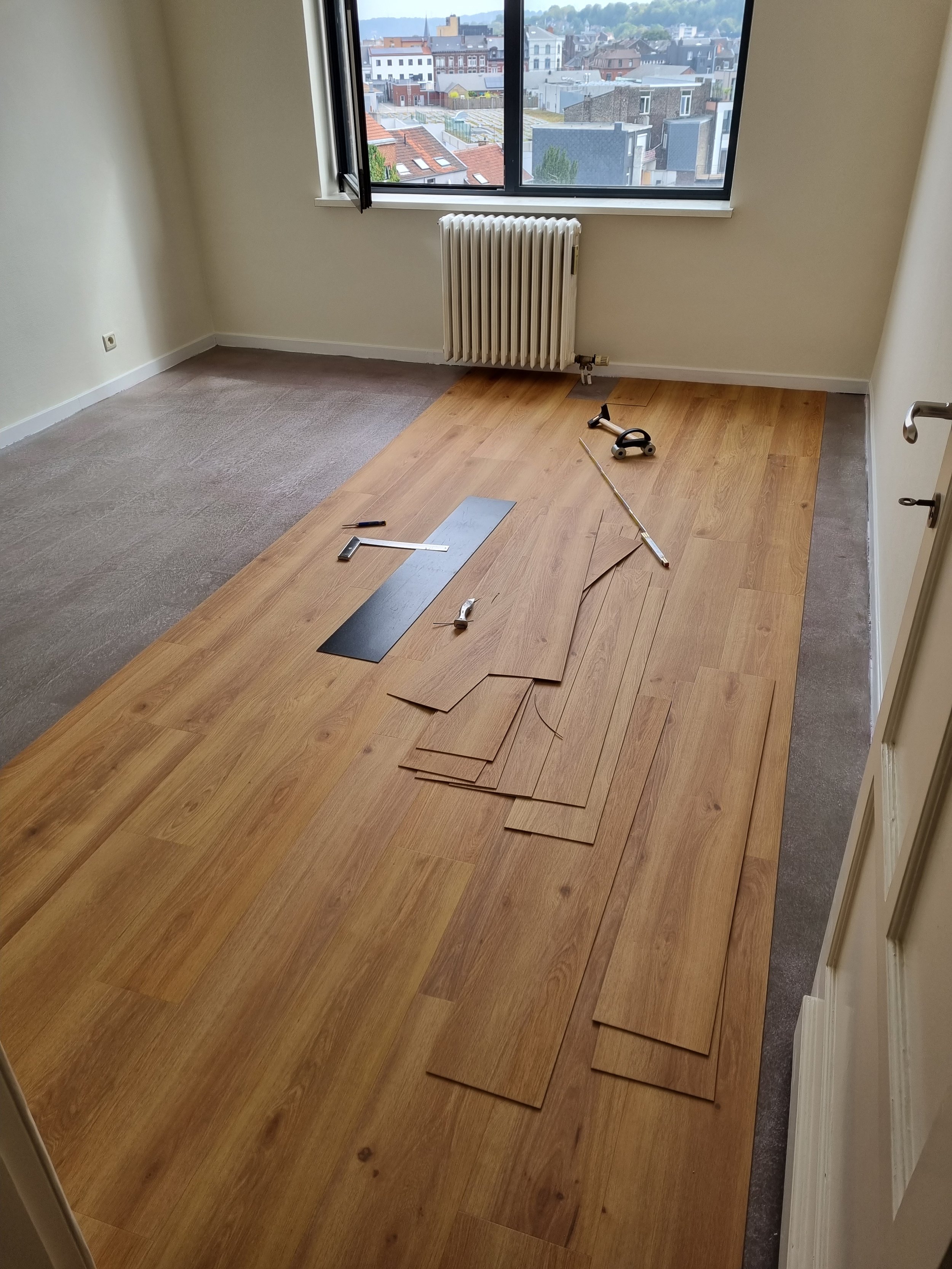 Une pièce en cours de rénovation avec des plaques de sol en bois clair posées sur une moitié du sol, laissant l'autre moitié à moitié recouverte de moquette. Il y a des outils pour la pose du parquet, notamment une règle, une scie, un marteau, et plusieurs plaques de bois. Une fenêtre donne sur une vue urbaine avec des bâtiments.