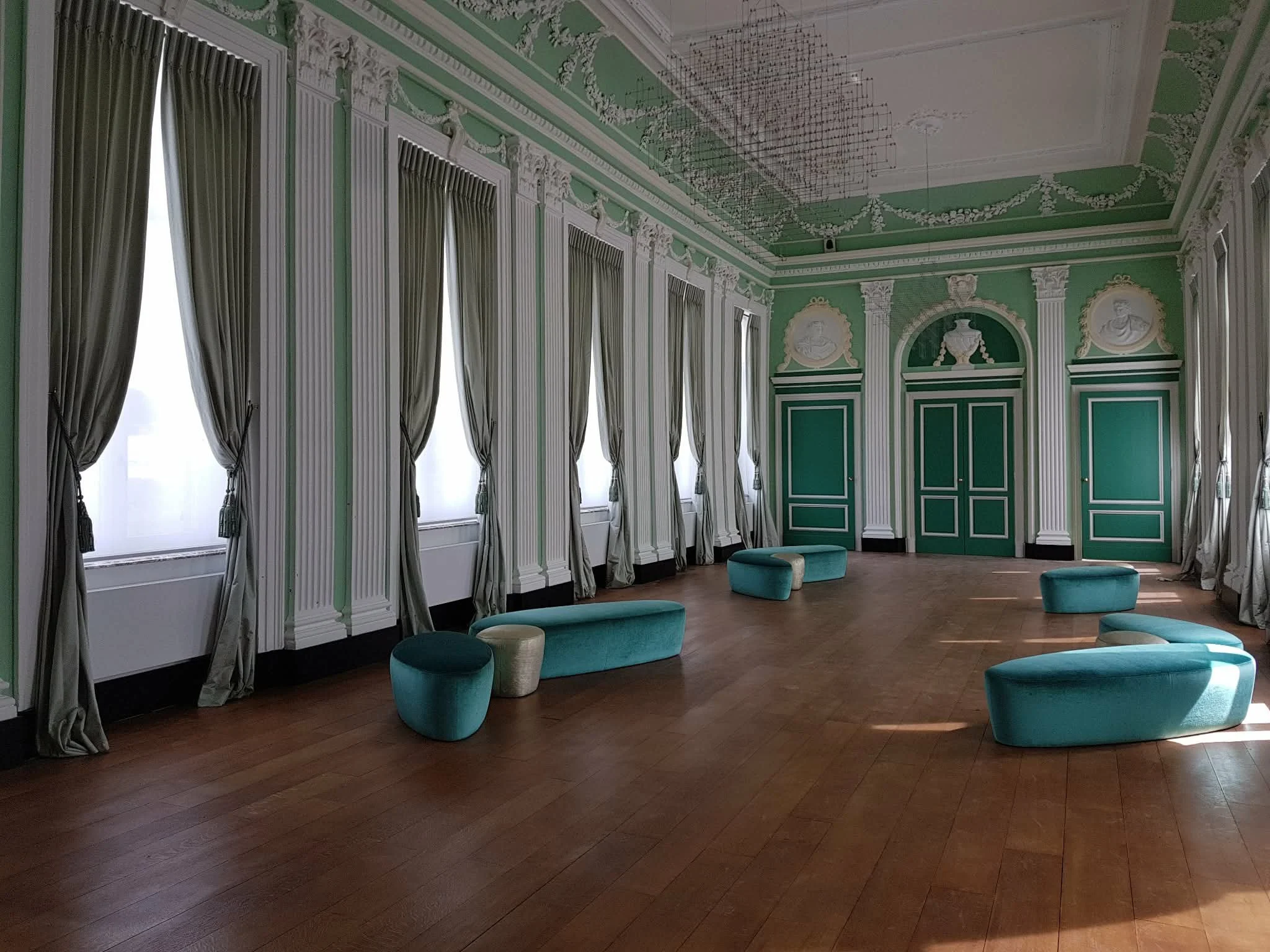 Salle de réception élégante avec murs verts, grands miroirs, rideaux gris, et fauteuils bleu turquoise en forme arrondie.