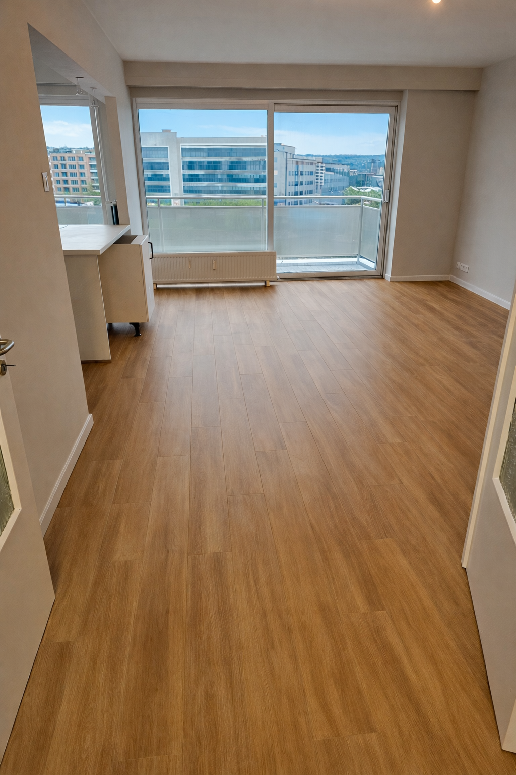Appartement vide avec parquet en bois clair, grande baie vitrée donne sur un balcon, vue d'immeubles modernes.