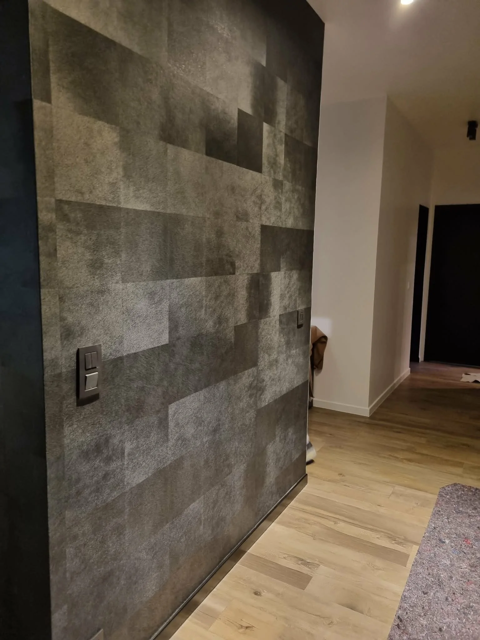 Un mur en papier peint gris foncé avec un motif géométrique dans une pièce intérieure, avec un interrupteur électrique visible.
