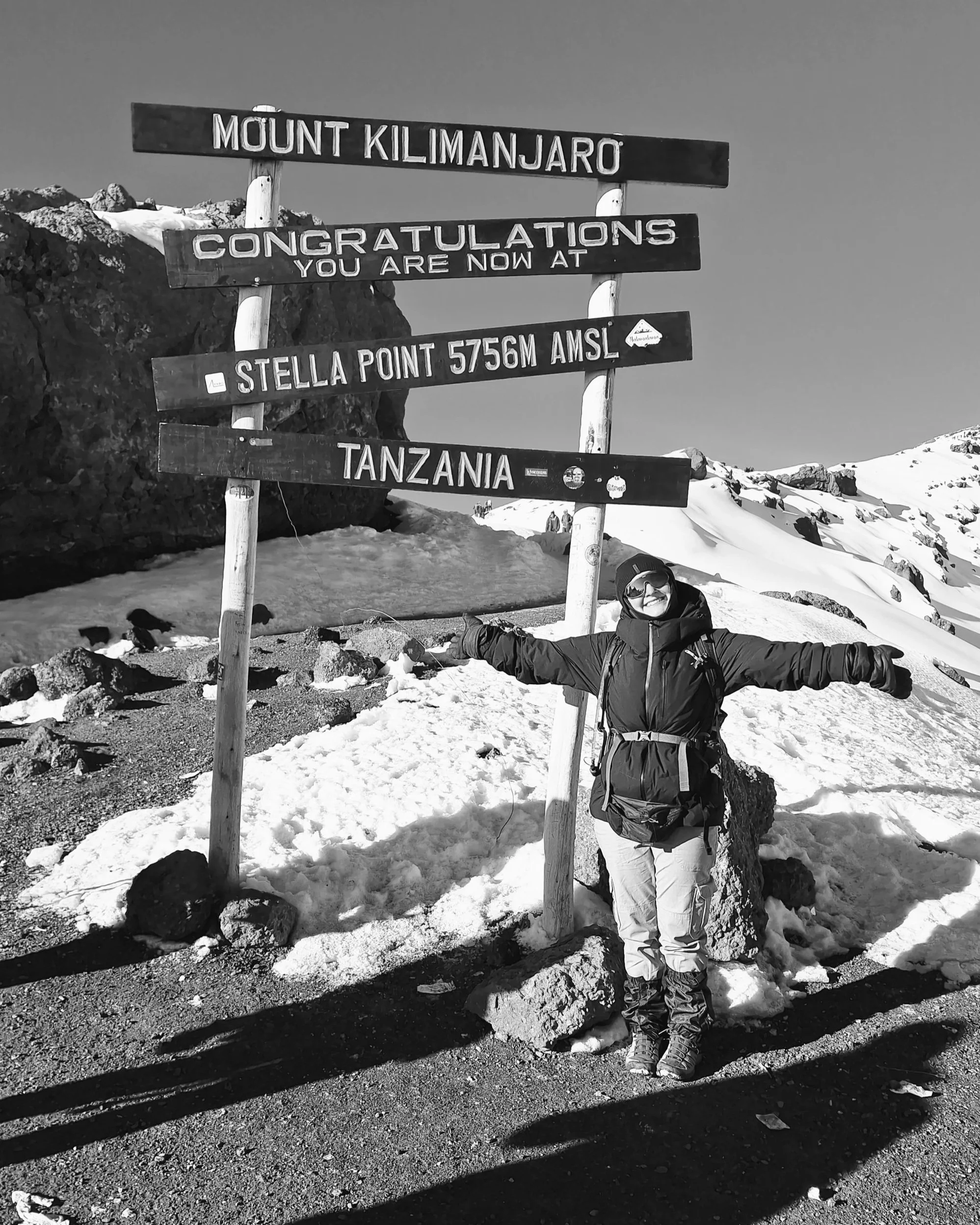 A person in winter gear standing on snow with arms outstretched, next to a sign that reads Mount Kilimanjaro, congratulations you are now at Stella Point 5756M AMSL, Tanzania, with snow-covered mountains and rocks in the background.