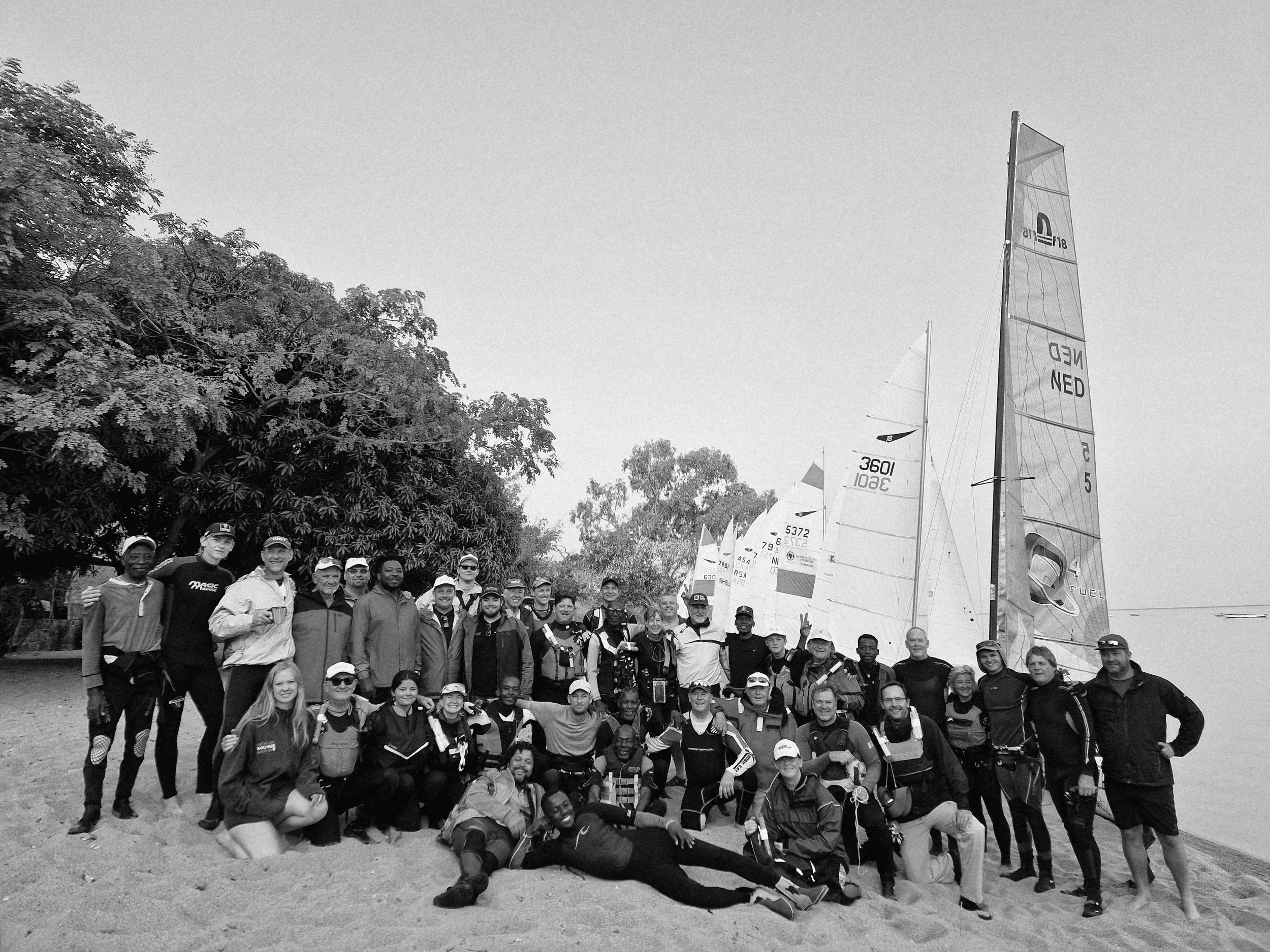 Group of people on a beach with sailboats in the background, some wearing life jackets and sailing gear, posing for a photo.