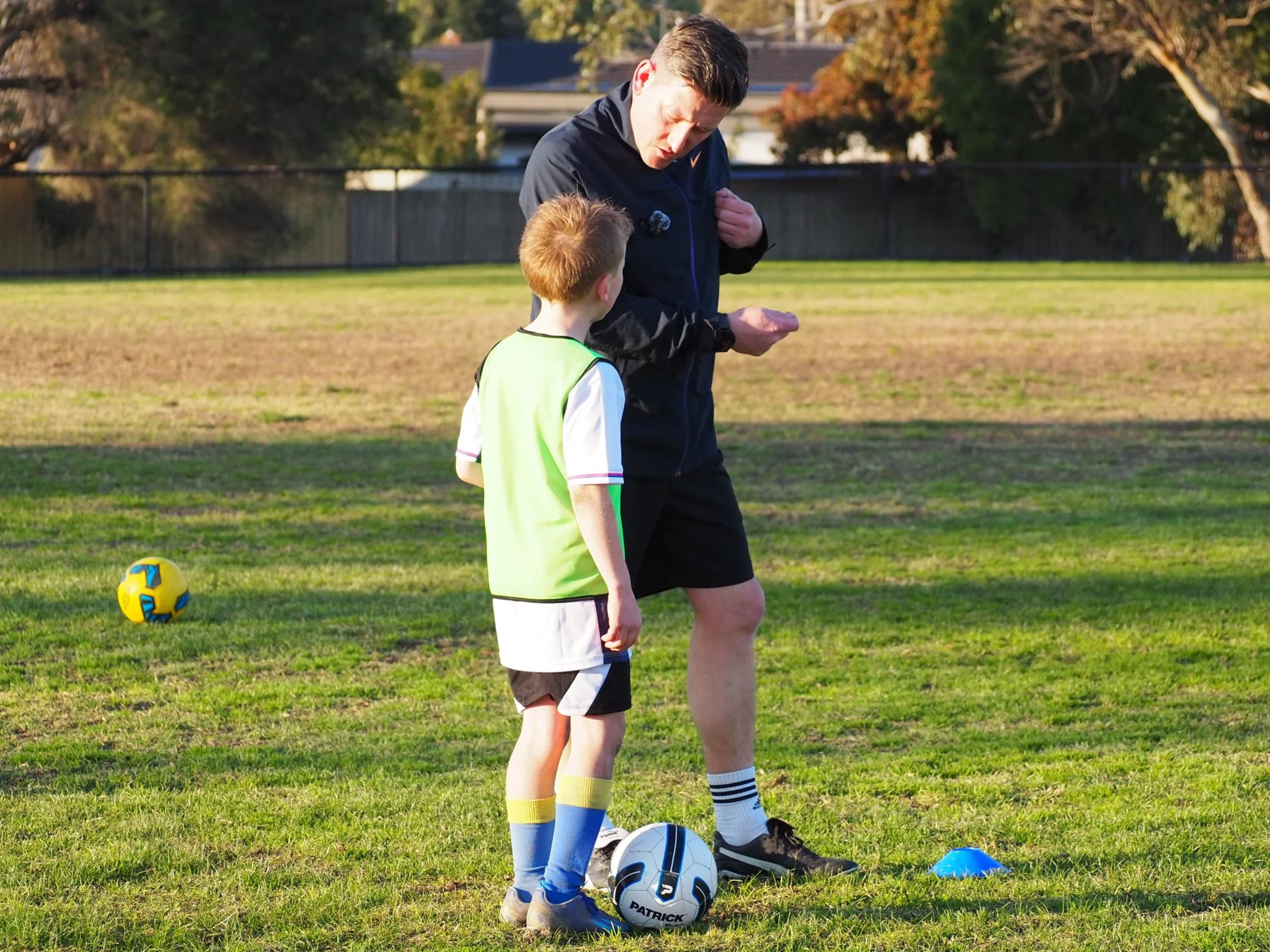 An adult male coach giving instructions to a young boy during a soccer practice on a field.