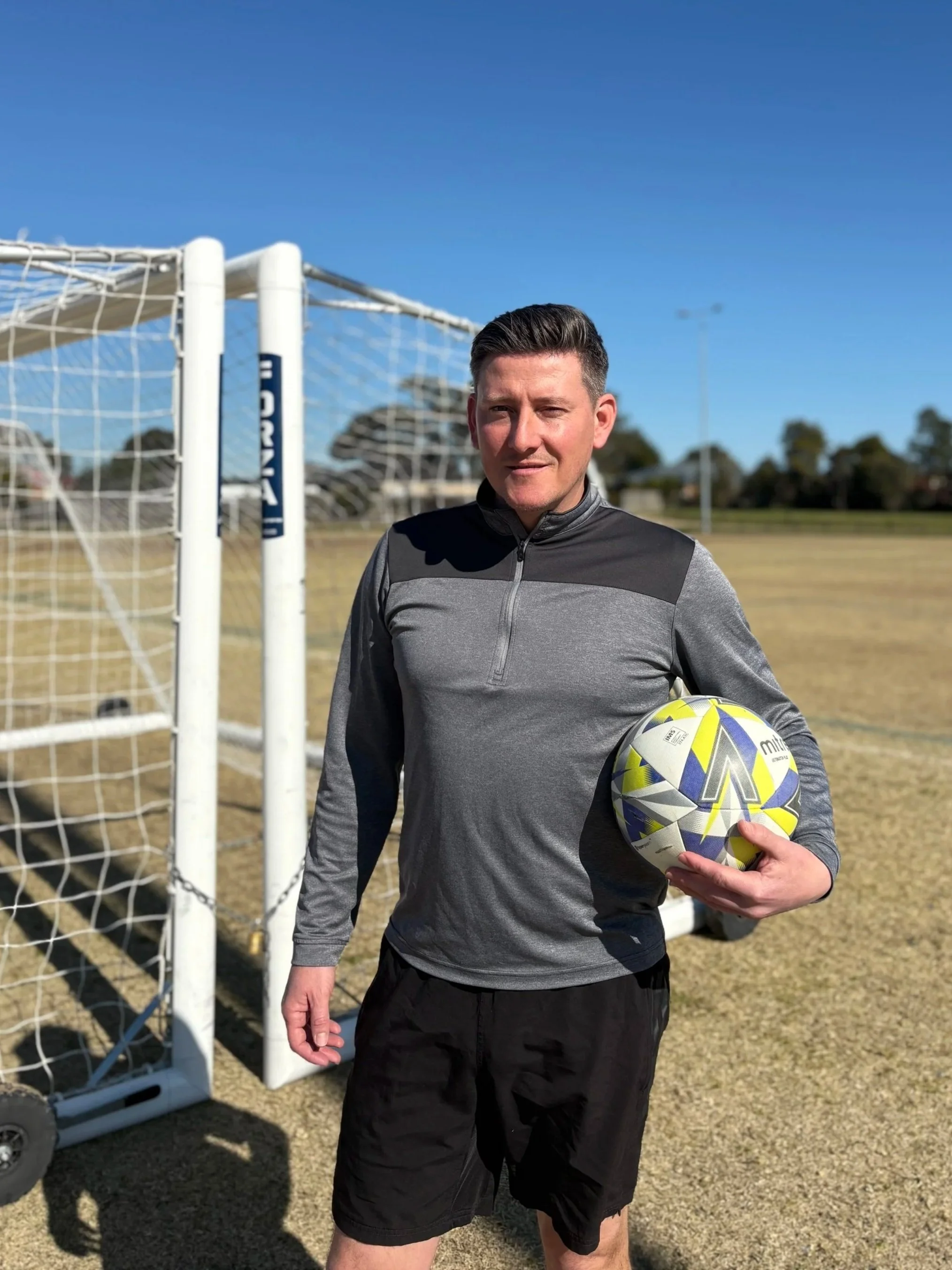 Soccer Coach, Mark Larner holding a soccer ball, with a soccer goal in the background, under a clear blue sky.