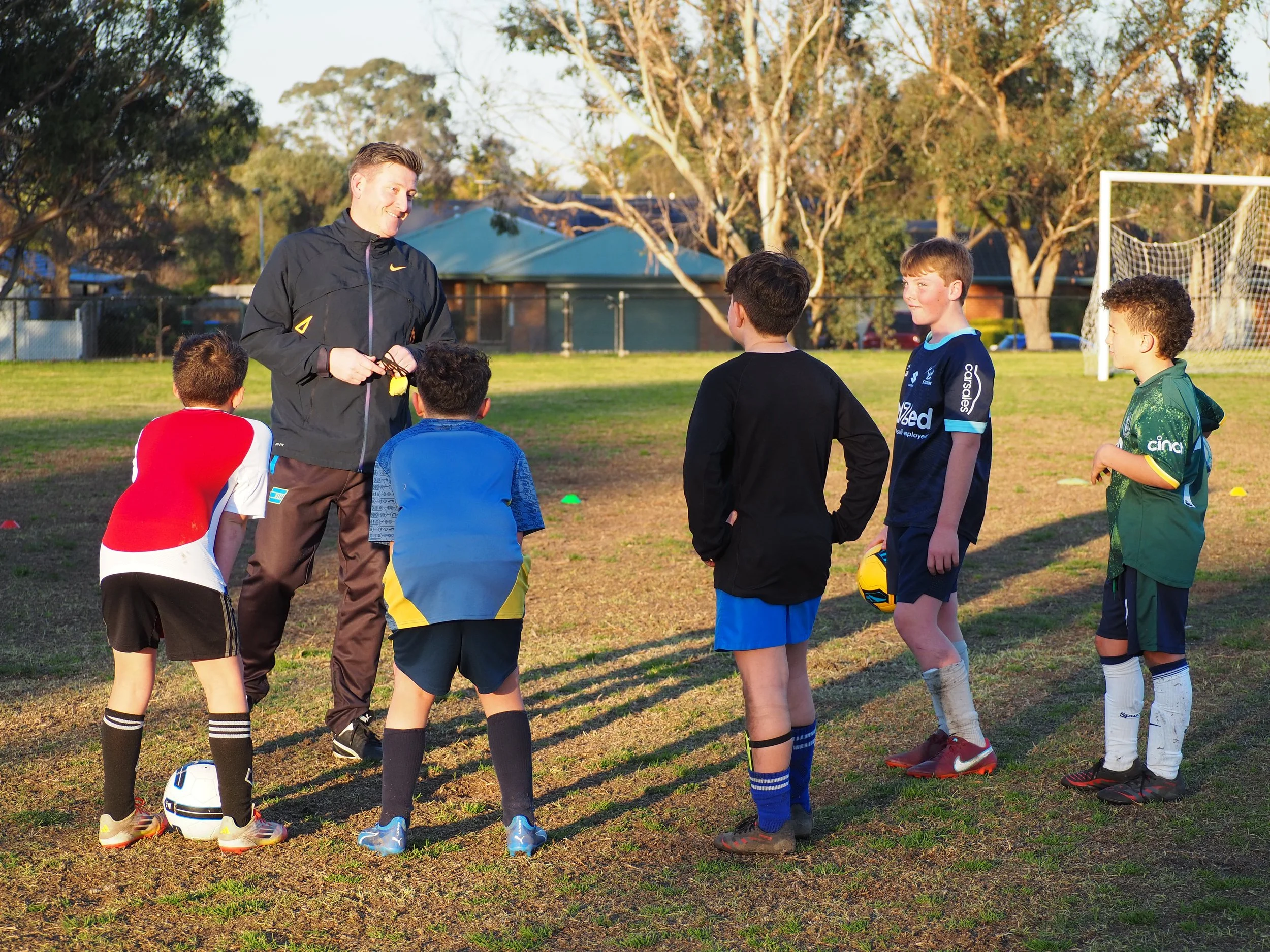 A coach giving instructions to a group of children on a soccer field with a goalpost in the background during sunset.