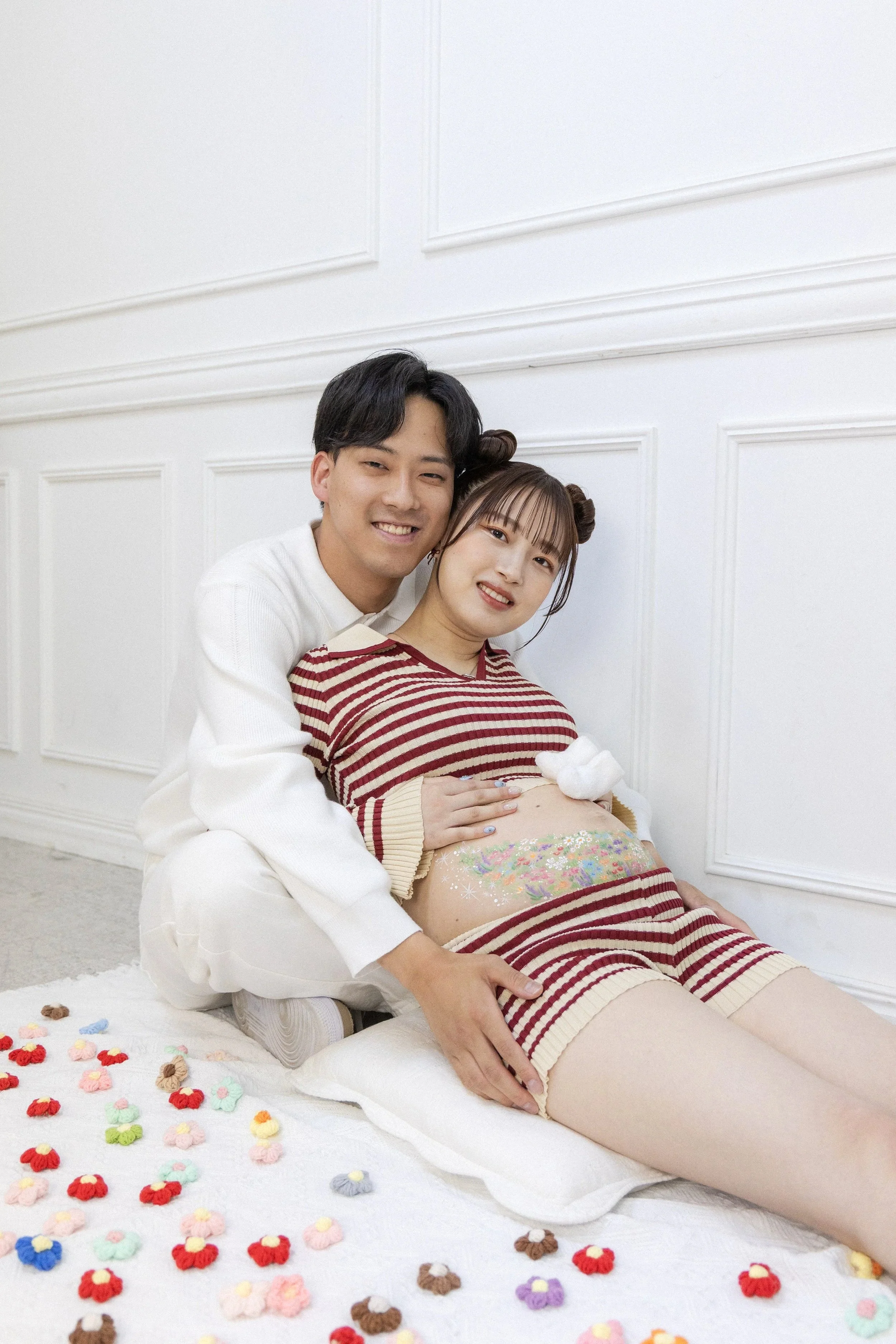 A couple sitting on a bed, with the woman leaning back against the man. The woman has her bare pregnant belly painted with colorful flowers and surrounded by small plush toys. They are smiling at the camera in a white room with decorative wall panels