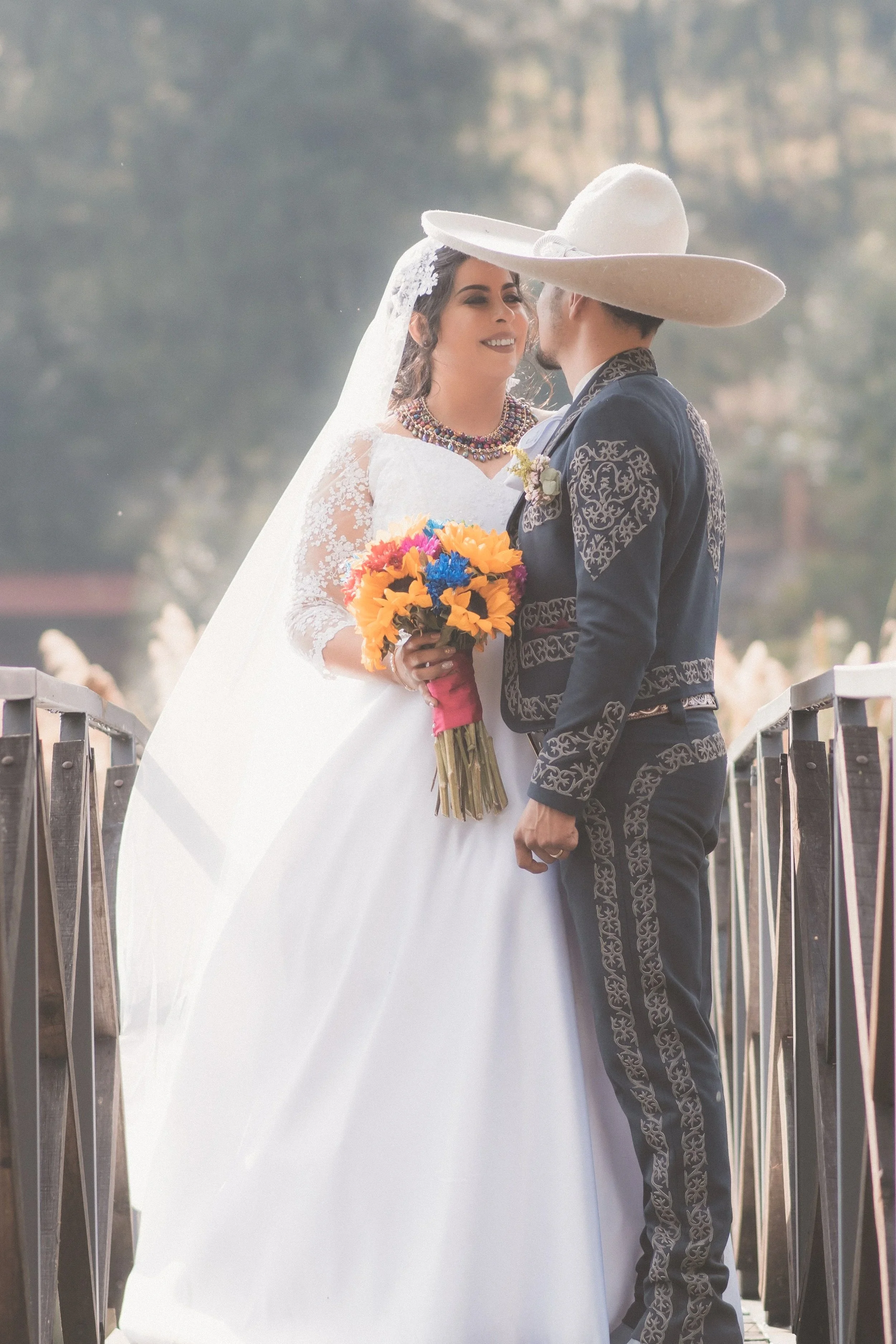 A bride and groom in traditional Mexican attire sharing a moment outdoors, with the bride holding a colorful bouquet and the groom wearing a large sombrero.