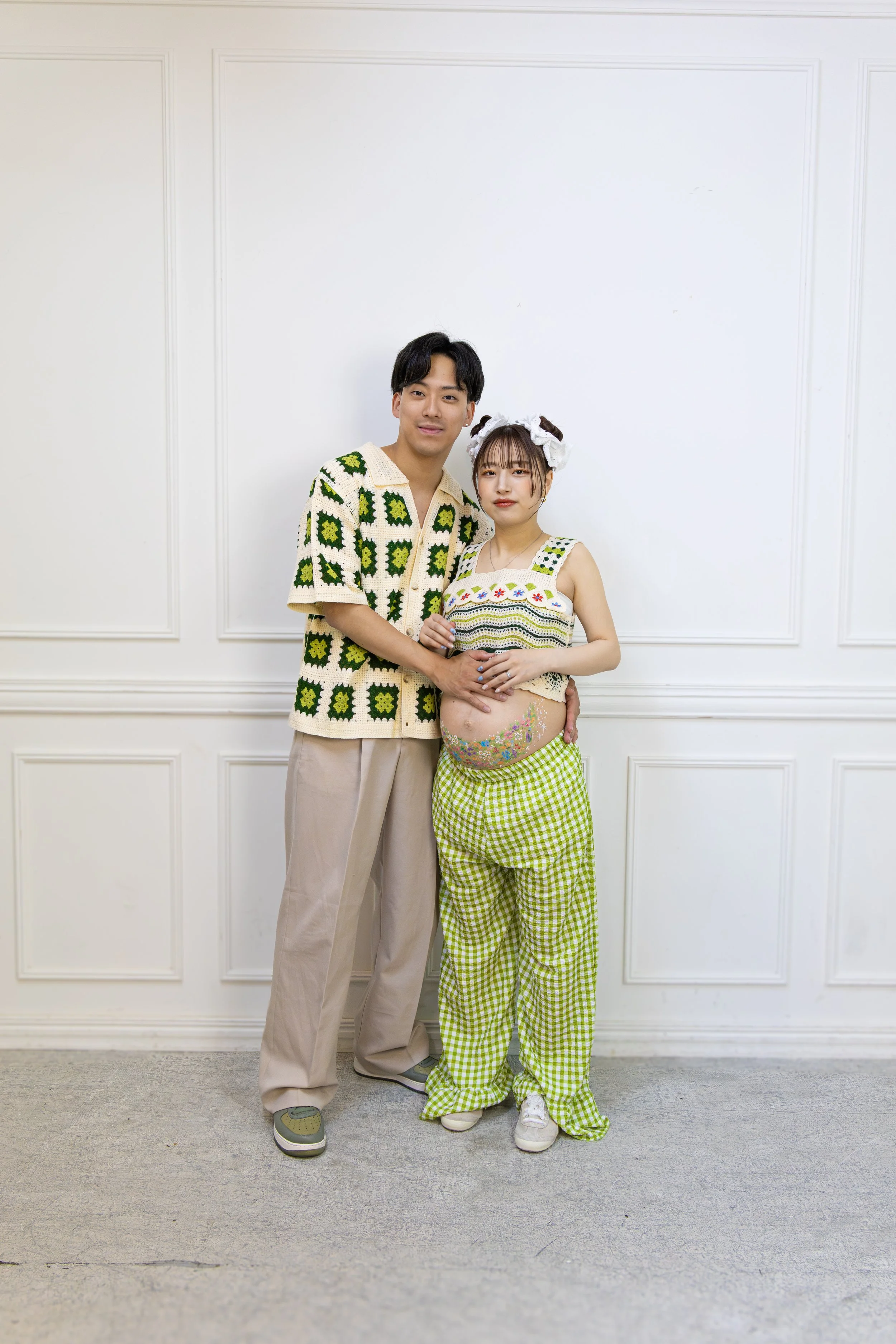 A couple standing together in front of a plain white wall. The woman has a pregnant belly adorned with colorful flowers, and she wears bright green checkered pants, a crocheted tops, and a floral headband. The man wears beige pants and a green and wh