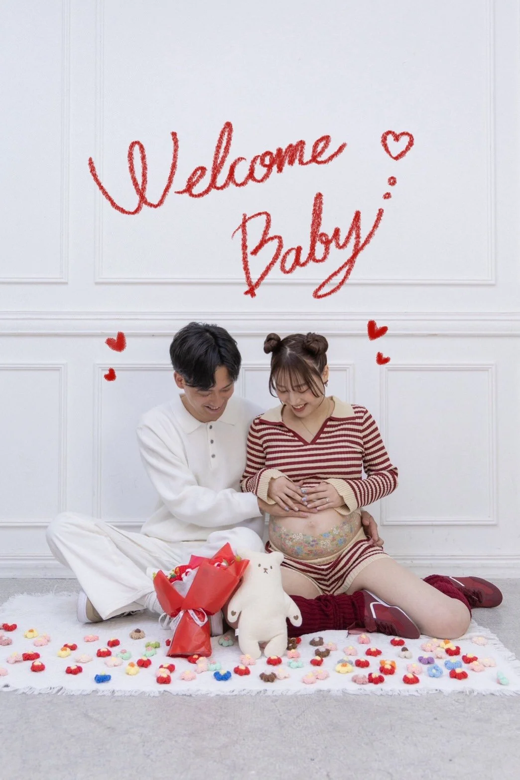 A happy couple celebrating a pregnancy in a room with a white wall and a sign that says 'Welcome Baby' in red. The woman is pregnant, wearing striped pajamas, and the man is sitting next to her. They are surrounded by colorful decorations, a bouquet,