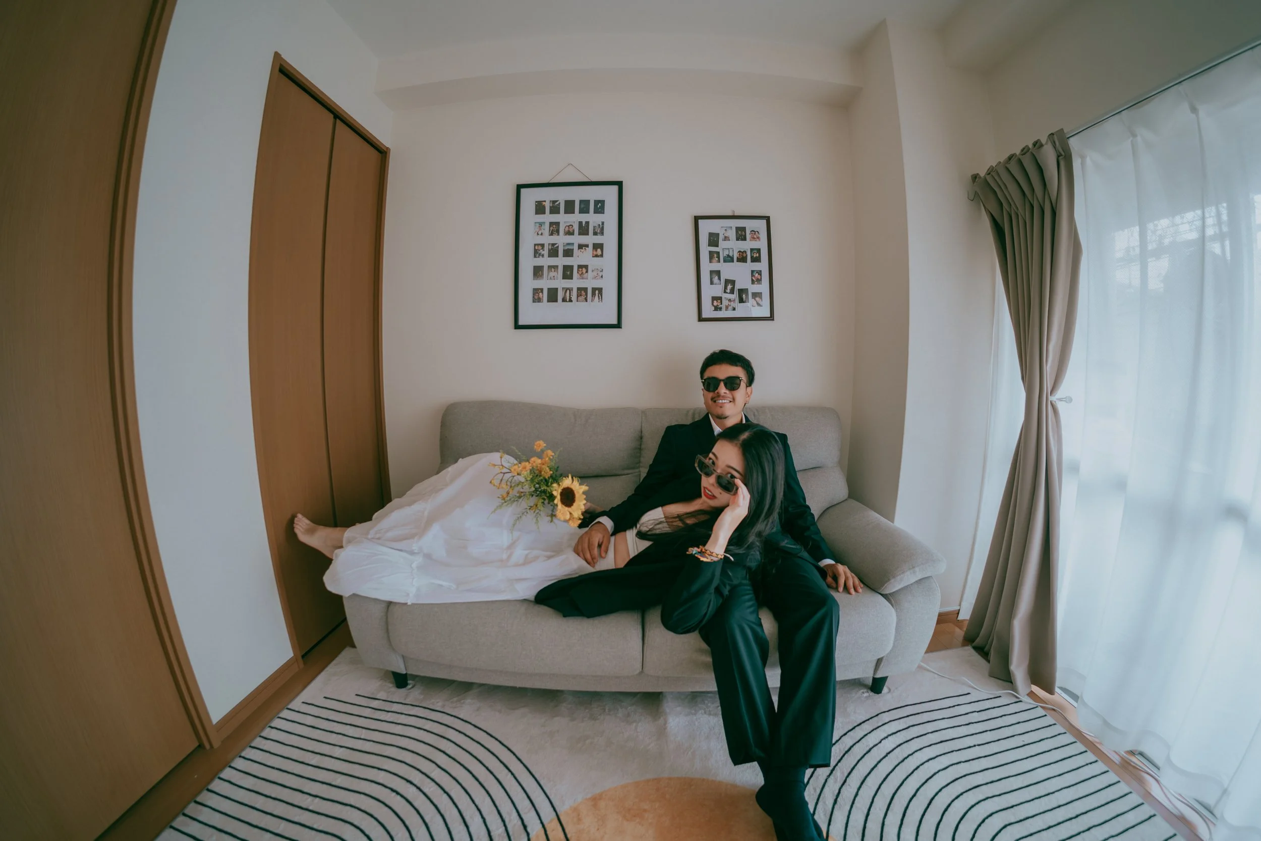 A man and woman in sunglasses sitting on a light gray sofa in a living room, with the woman holding a bouquet of flowers, and a window with curtains on the right side.