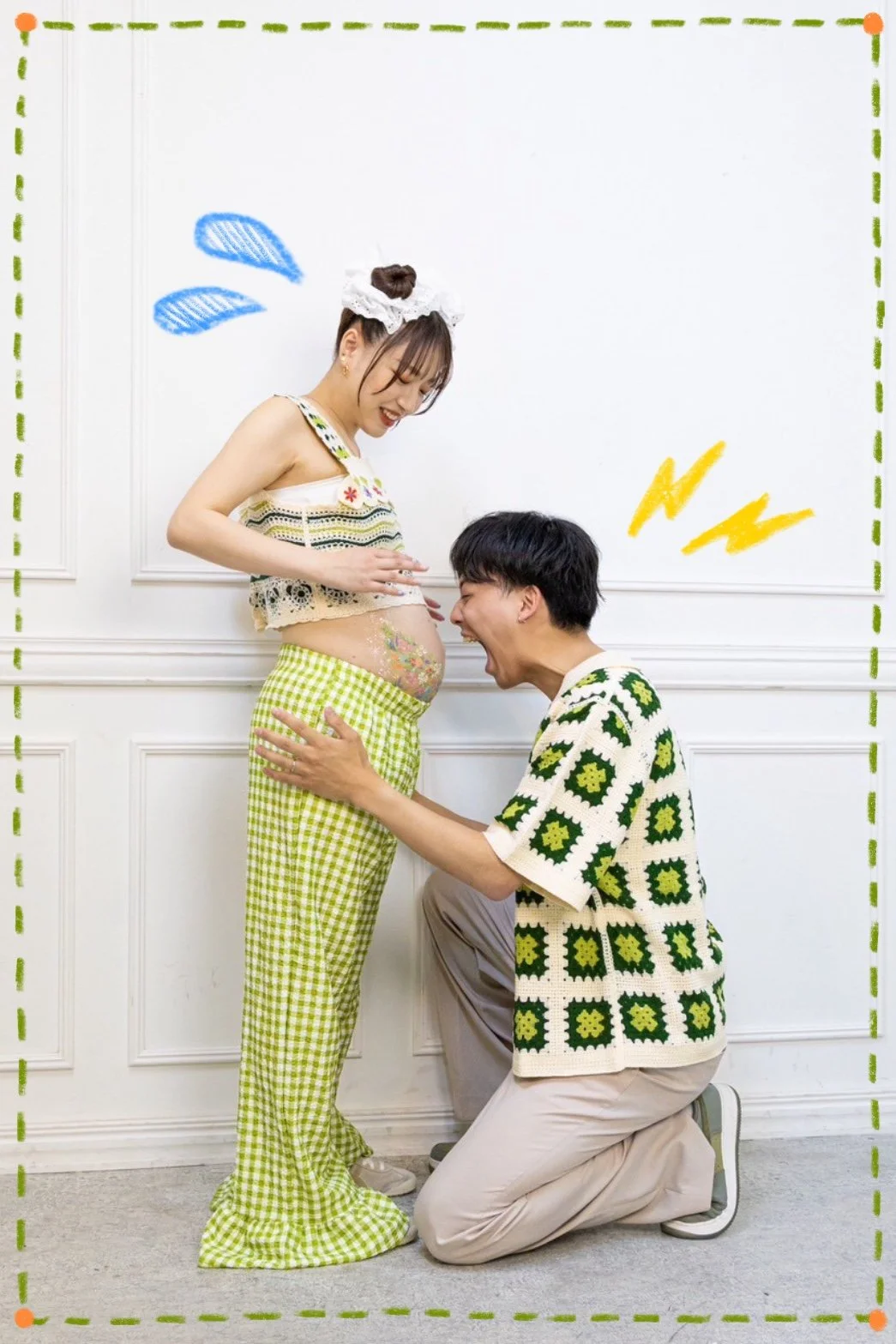 A pregnant woman and a man are interacting playfully indoors against a white wall. The woman is standing, wearing green and white checked pants and a lace top, with colorful tattoos on her belly. The man is kneeling in front of her, wearing a pattern
