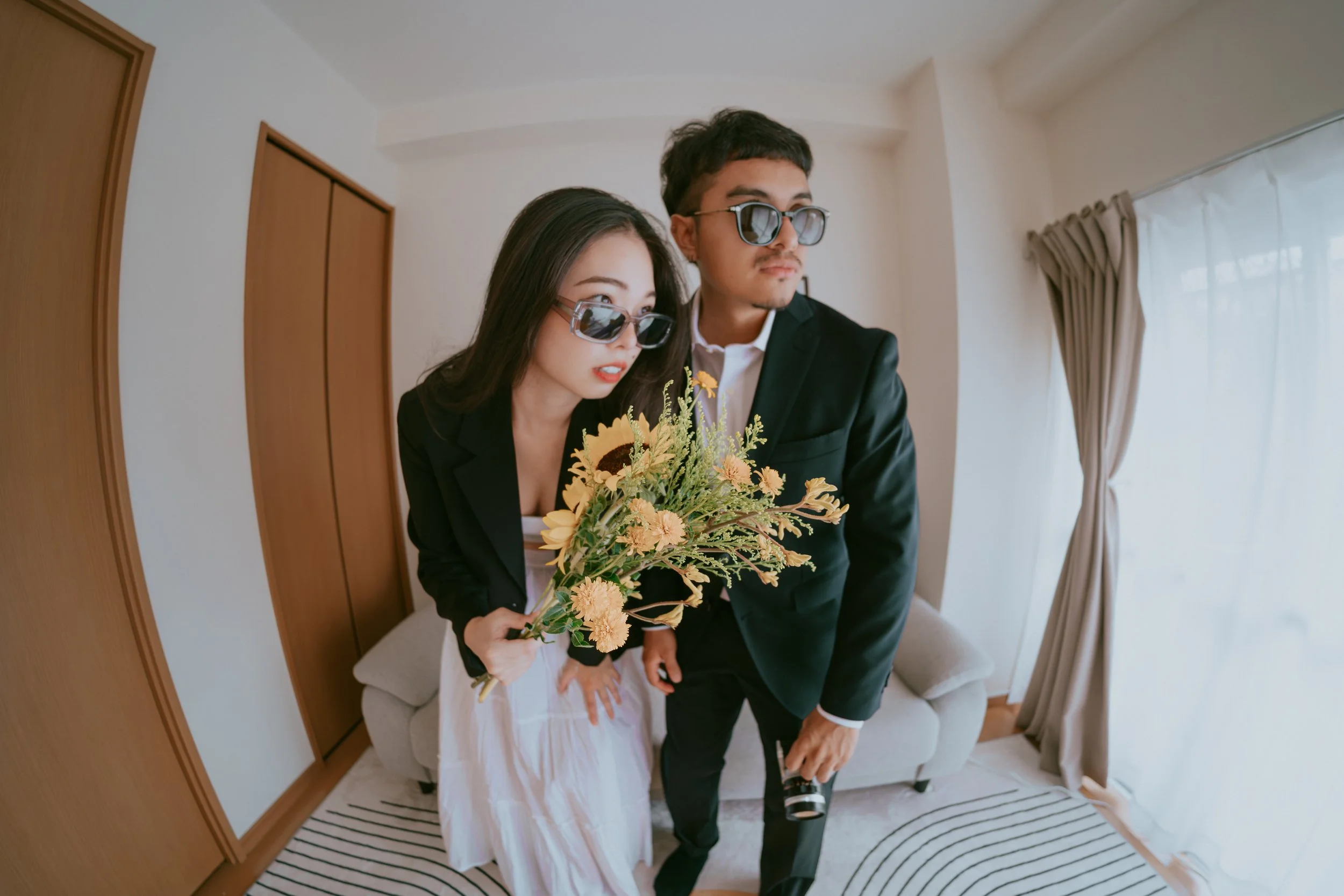 A young woman and man, both wearing sunglasses and dressed in black suits, stand in a room with white walls and a large window with curtains. The woman holds a bouquet of sunflowers and other yellow flowers, and the man holds a camera in his right hand.